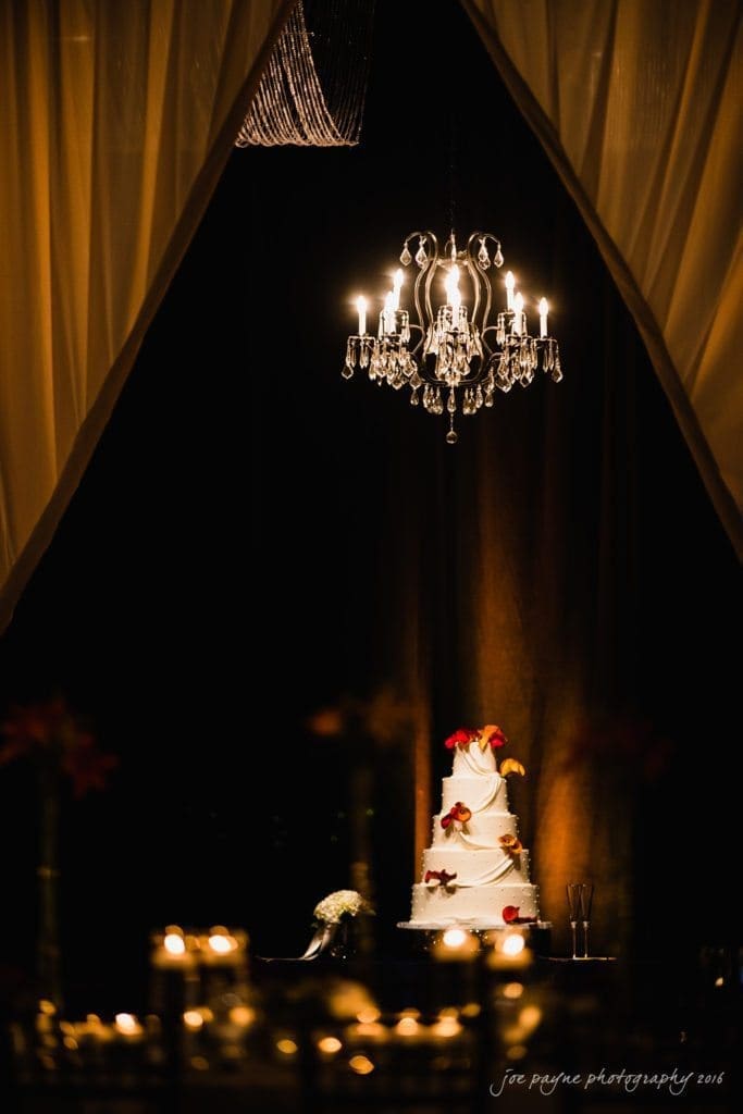 winston-salem nc wedding photography - toni & kevin A tall, white wedding cake adorned with red and orange flowers sits under a lit chandelier. The cake is displayed in a dimly lit room framed by sheer curtains, creating an elegant and intimate atmosphere.