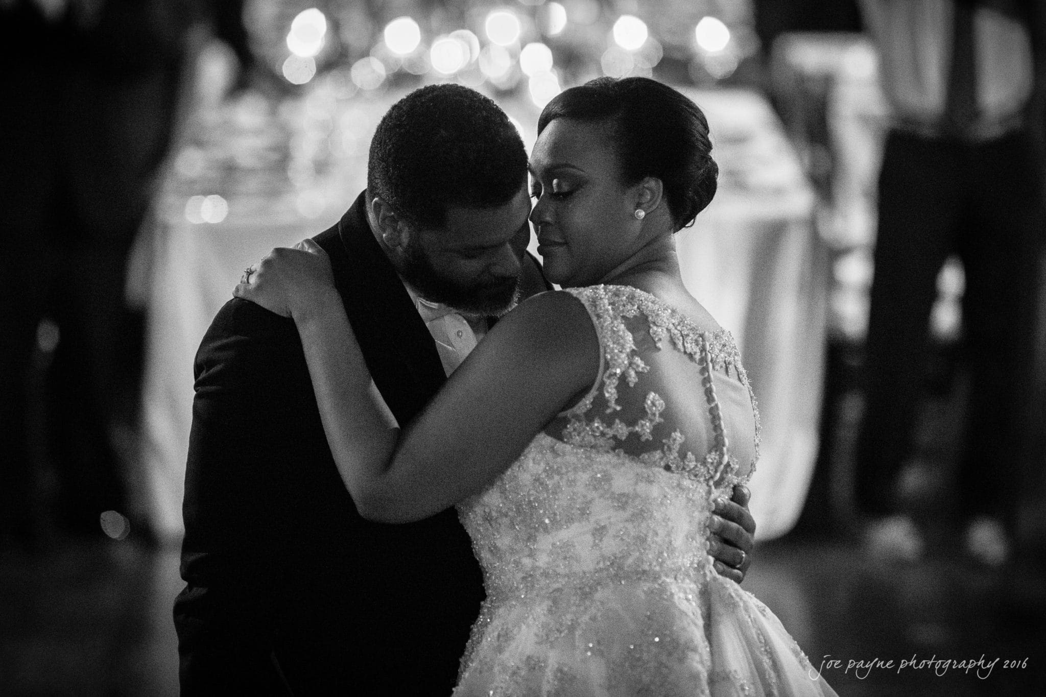 winston-salem nc wedding photography - toni & kevin A couple shares a tender dance at a wedding. The man is wearing a dark suit and the woman is in a detailed, sleeveless wedding dress. The background is softly blurred, with glowing lights creating a romantic atmosphere.