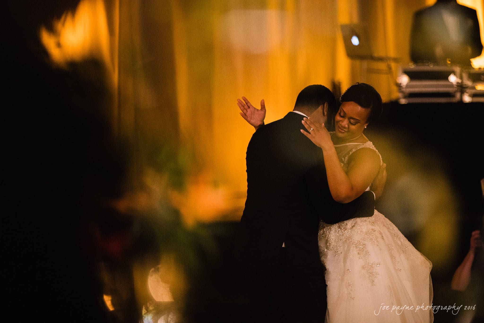 winston-salem nc wedding photography - toni & kevin A couple shares a warm embrace during a slow dance at a wedding reception. The bride wears a white gown, and the groom is in a black suit. They are surrounded by soft, warm golden lighting, creating an intimate and romantic atmosphere.