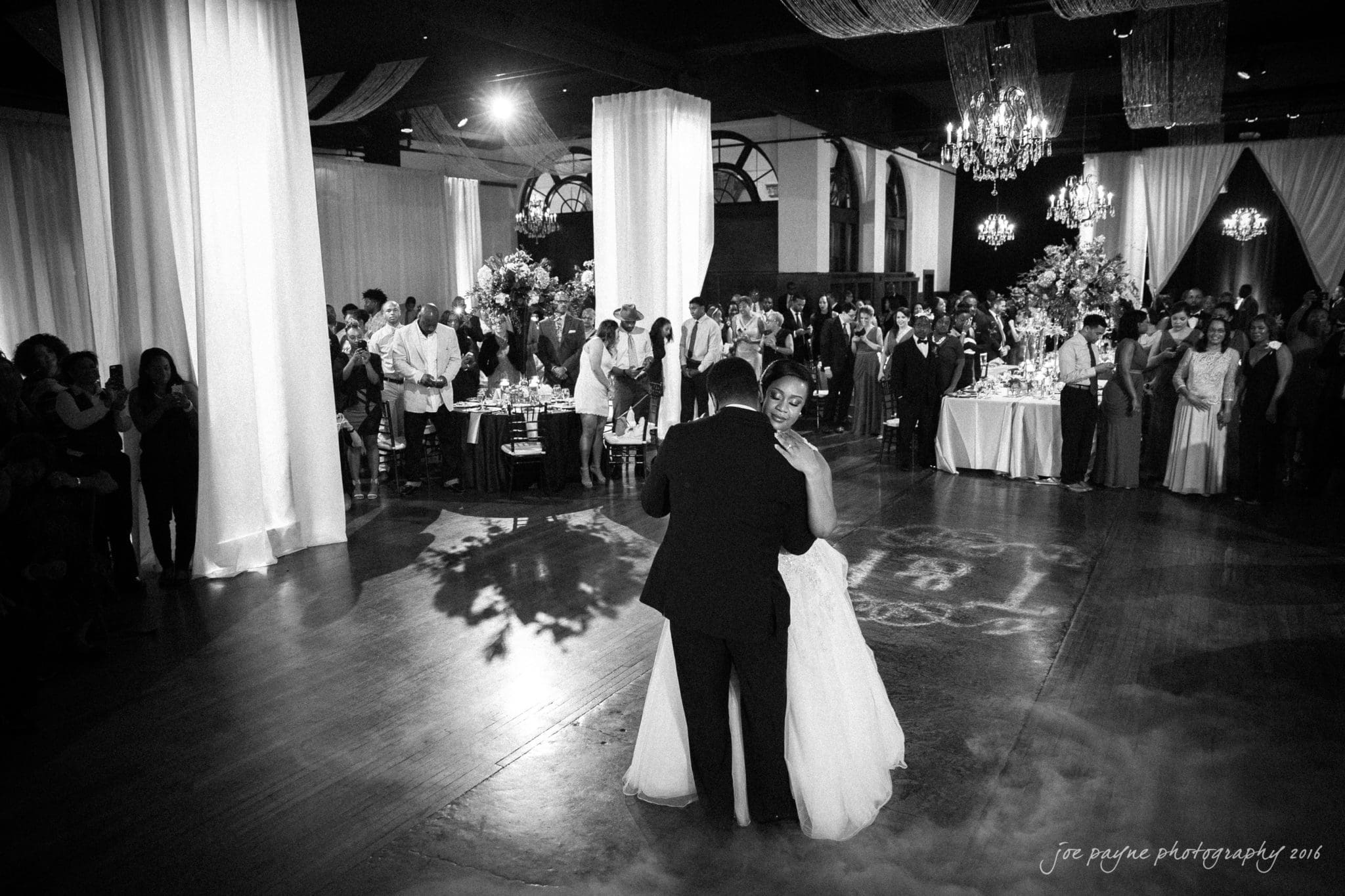 winston-salem nc wedding photography - toni & kevin A couple shares their first dance at a wedding reception in a dimly lit ballroom. Guests stand around watching, with elegantly decorated tables and floral arrangements in the background. A chandelier hangs from the ceiling, adding to the romantic ambiance.