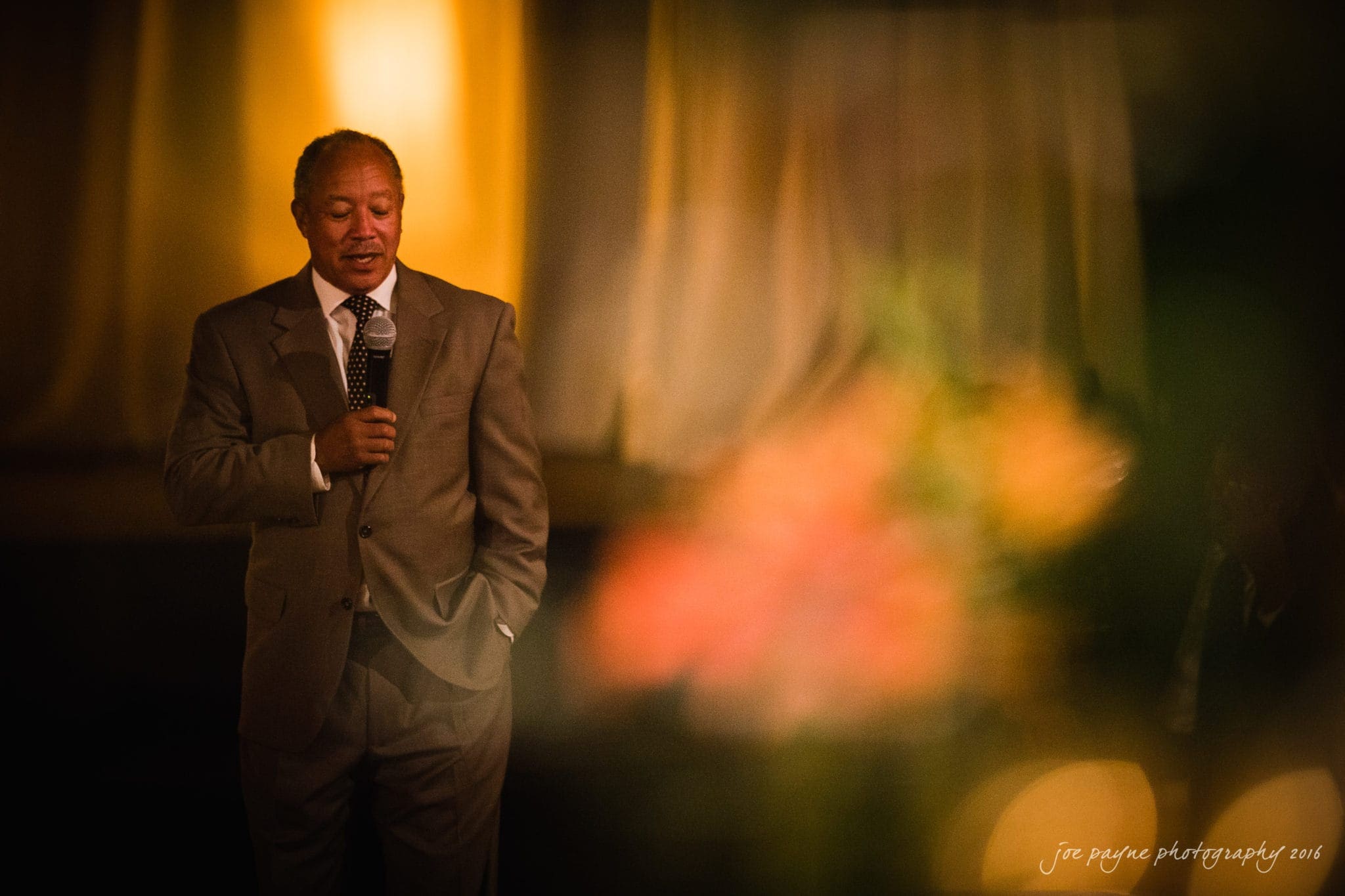 winston-salem nc wedding photography - toni & kevin A man in a brown suit holds a microphone while speaking softly, standing against a blurred, warm-toned background.