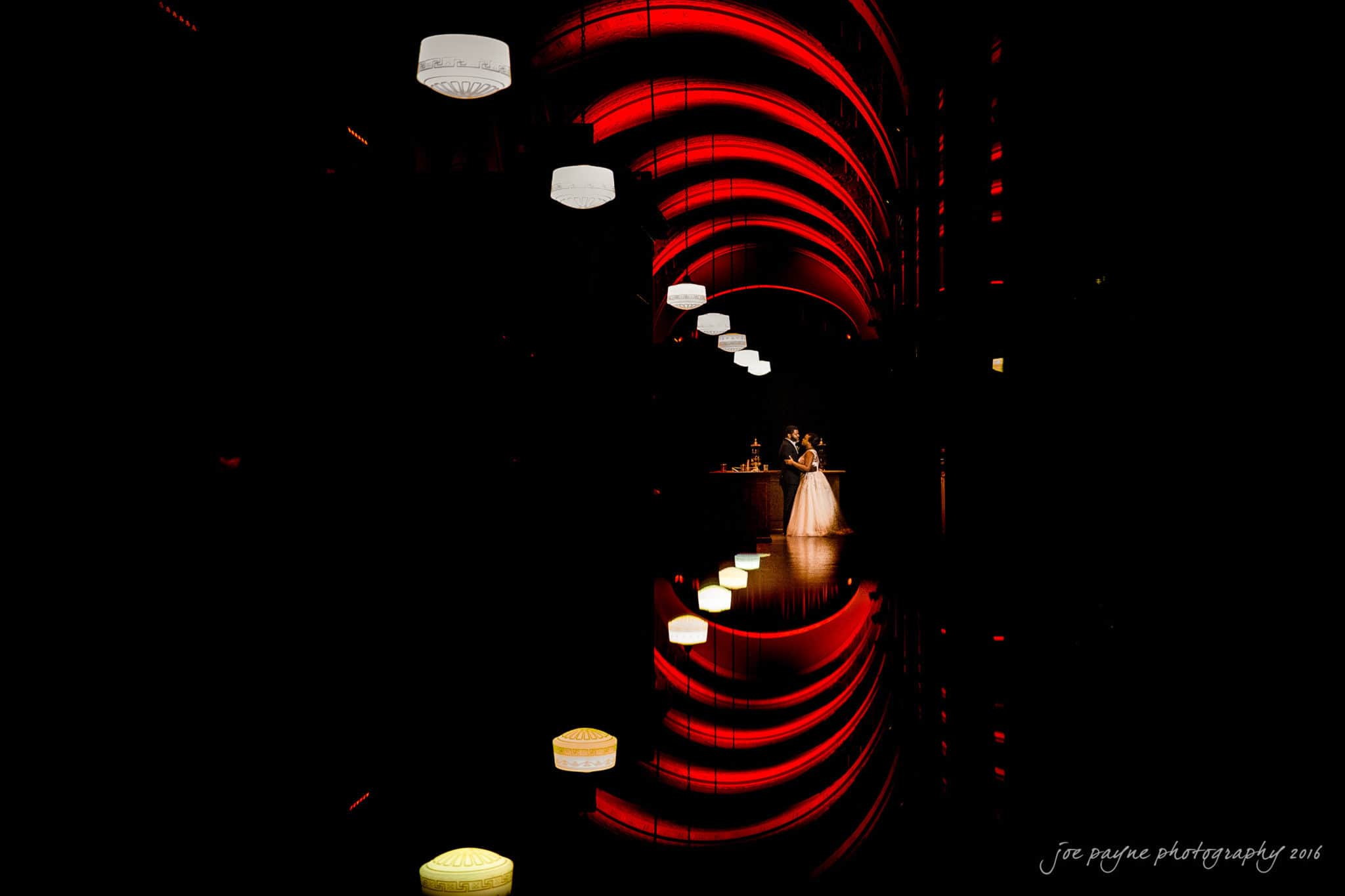 winston-salem nc wedding photography - toni & kevin A bride and groom share a moment on a dark balcony under arched red lighting, with their reflection visible below. Bright white and red lights line the path, adding a dramatic effect to the romantic scene.