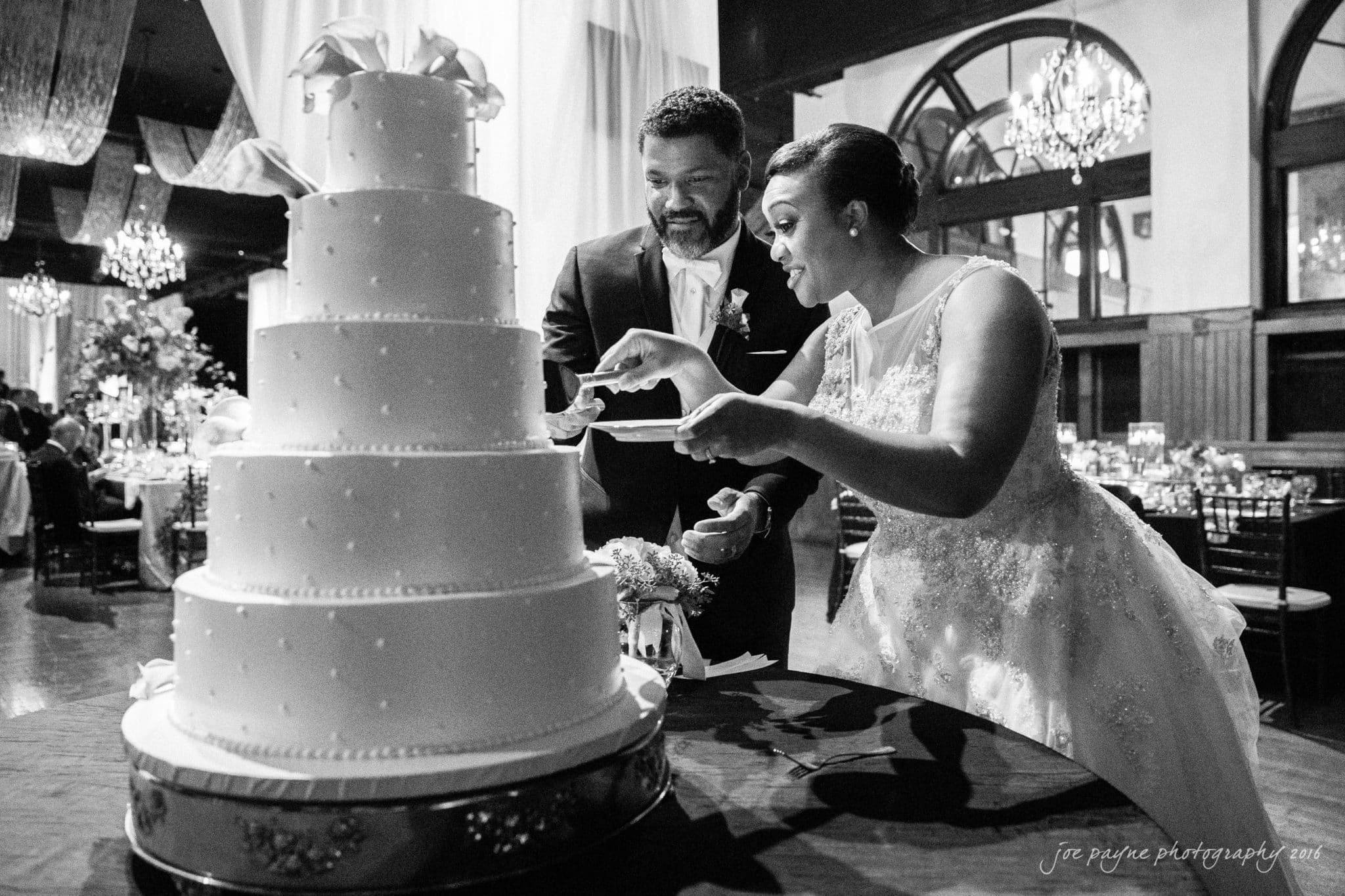 winston-salem nc wedding photography - toni & kevin A joyful couple in wedding attire are about to cut a three-tiered wedding cake at a reception. The elegant room is decorated with chandeliers and draped curtains, creating a festive atmosphere.