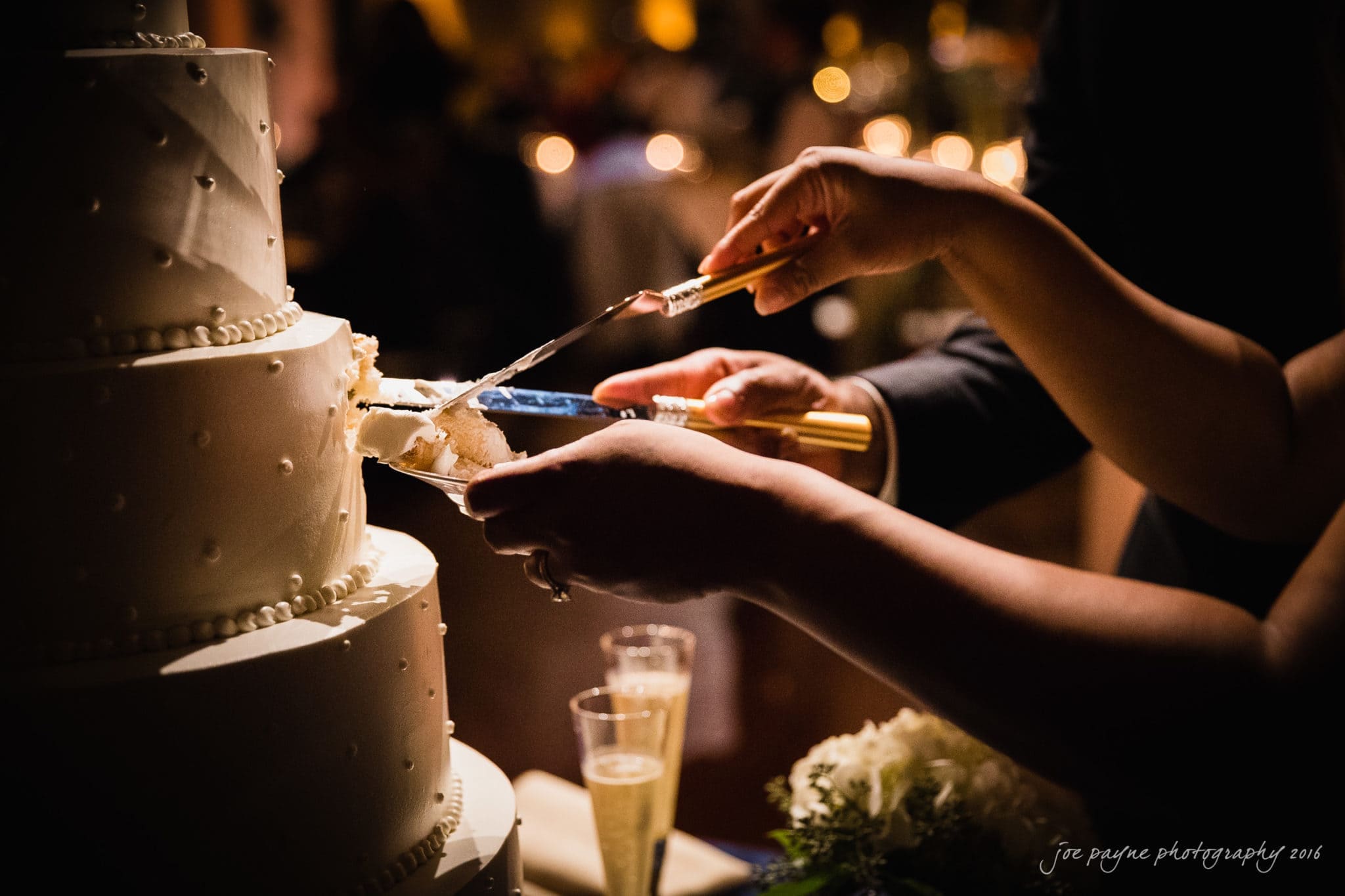 winston-salem nc wedding photography - toni & kevin A close-up of two people cutting a tiered wedding cake adorned with small decorative dots. The scene is warmly lit, creating a celebratory atmosphere. Two glasses of champagne and floral arrangements are visible in the foreground.