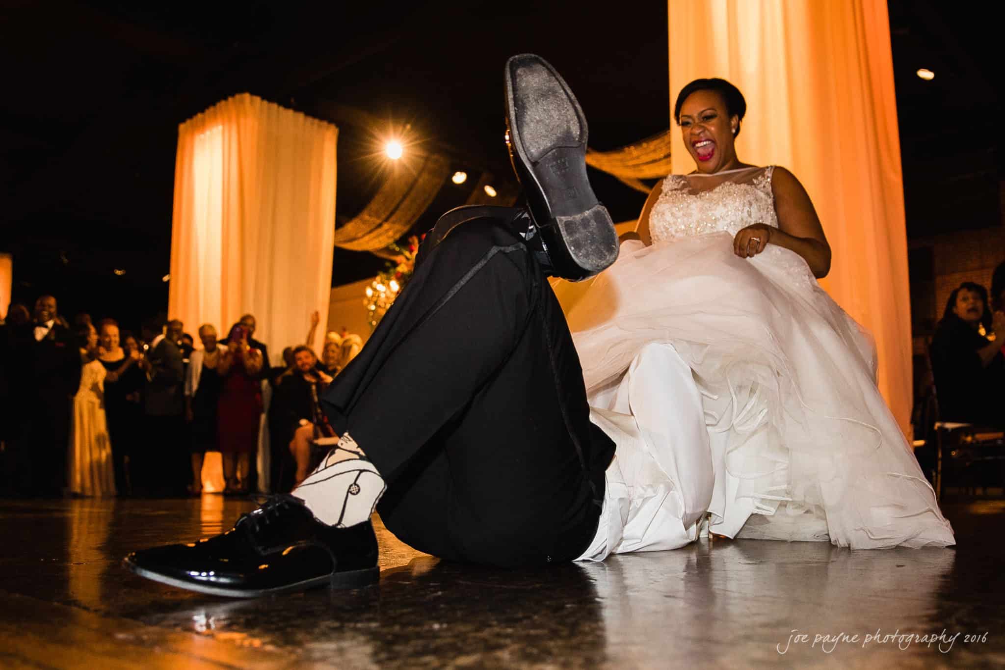 winston-salem nc wedding photography - toni & kevin A bride laughs joyfully as the groom, lying on the floor, raises his shoe-clad foot playfully. Guests in the background cheer and clap. The room is warmly lit with peach-colored drapery.