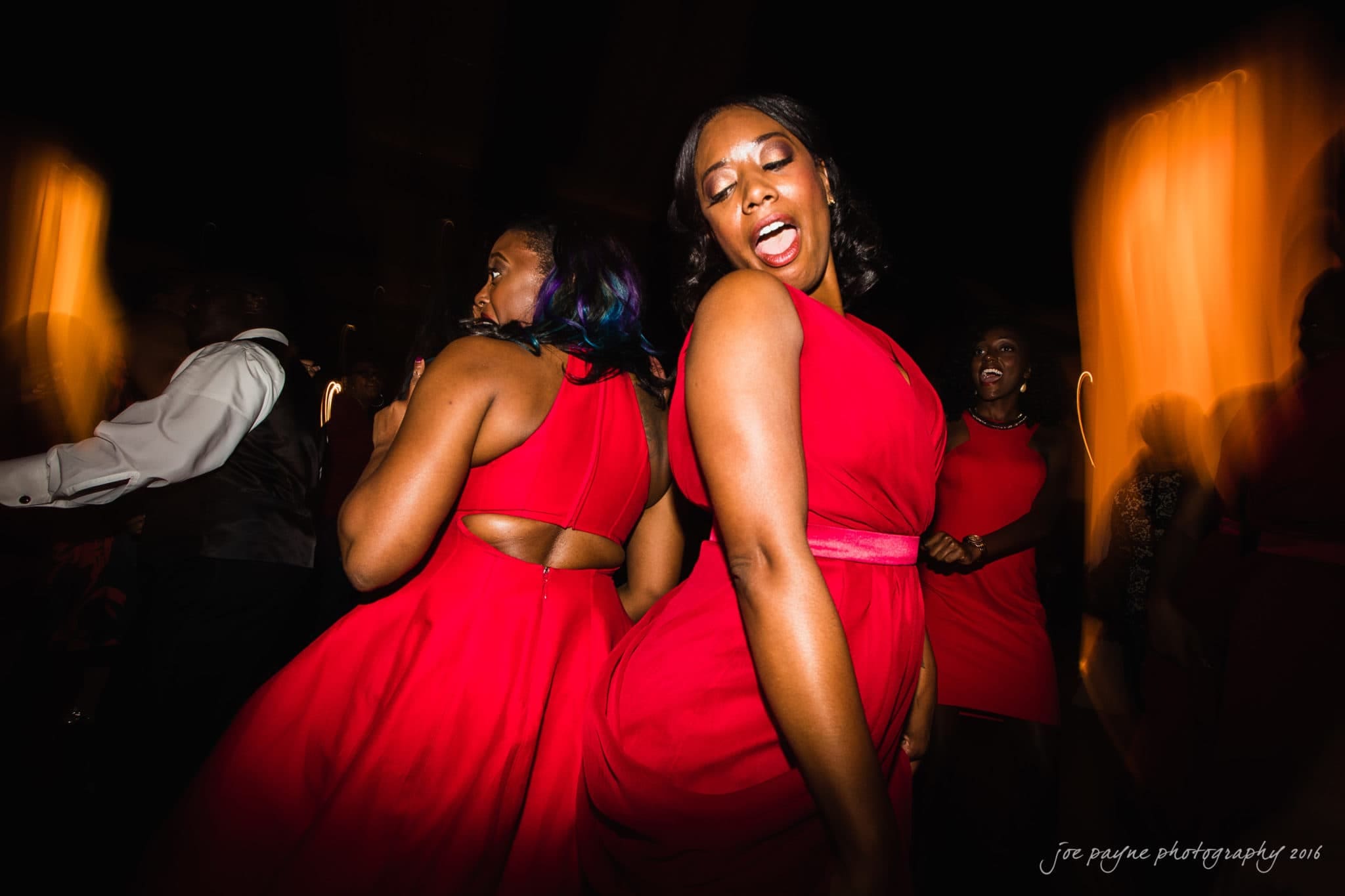 winston-salem nc wedding photography - toni & kevin Two women in red dresses dance energetically at a dimly lit event. They are smiling and appear to be enjoying themselves, with blurred motion effects highlighting the lively atmosphere.