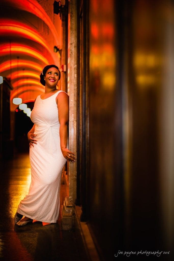 winston-salem nc wedding photography - toni & kevin A woman in a sleeveless white gown poses elegantly against a wall in a warmly lit hallway with arched ceilings. Her hand rests on her hip, and she gazes to the side with a smile. The lighting creates a soft, glowing atmosphere.