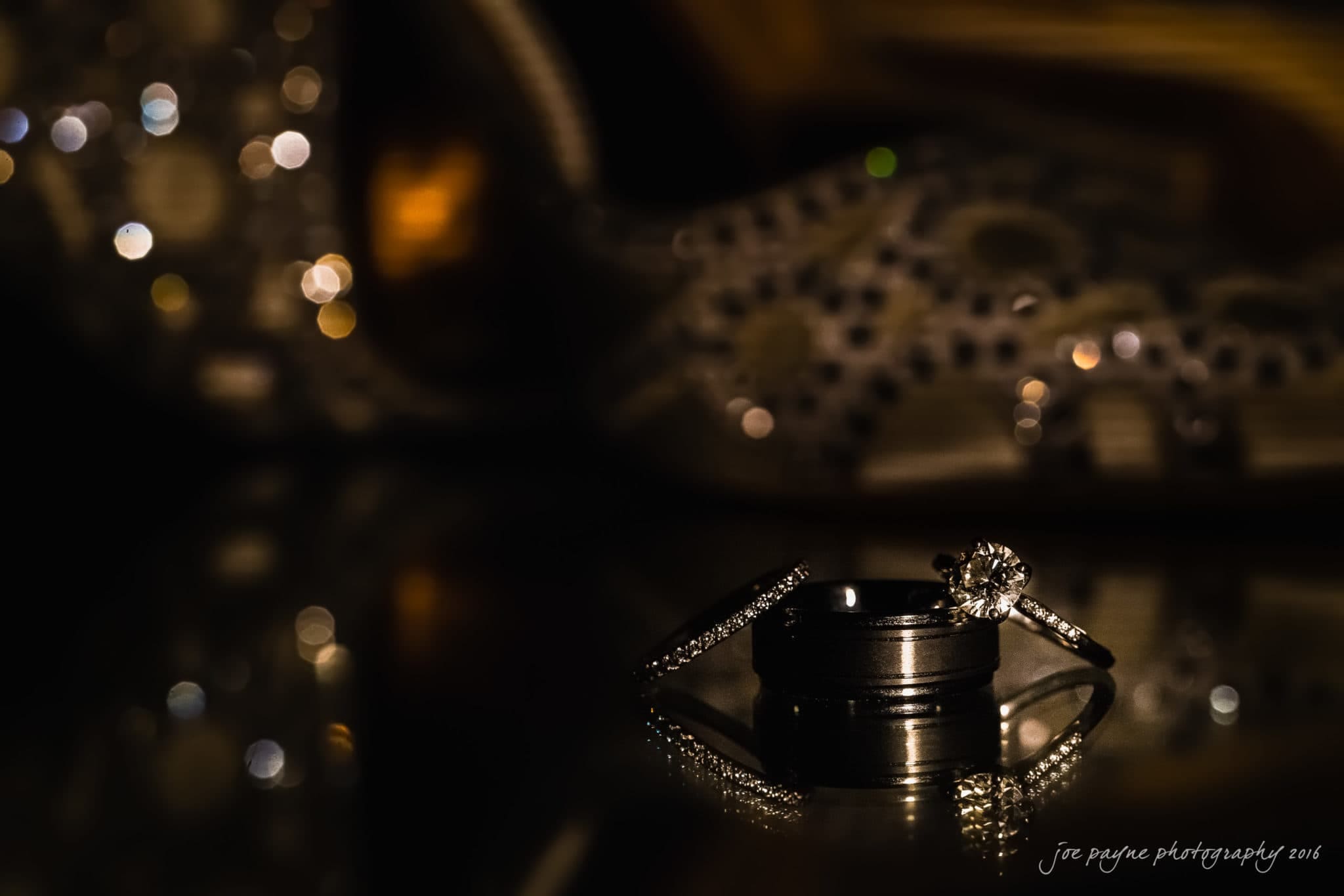 winston-salem nc wedding photography - toni & kevin Close-up of two wedding bands and a diamond engagement ring on a reflective surface, with dim lighting highlighting the jewelry against a blurred background of intricate patterns and soft bokeh lights.