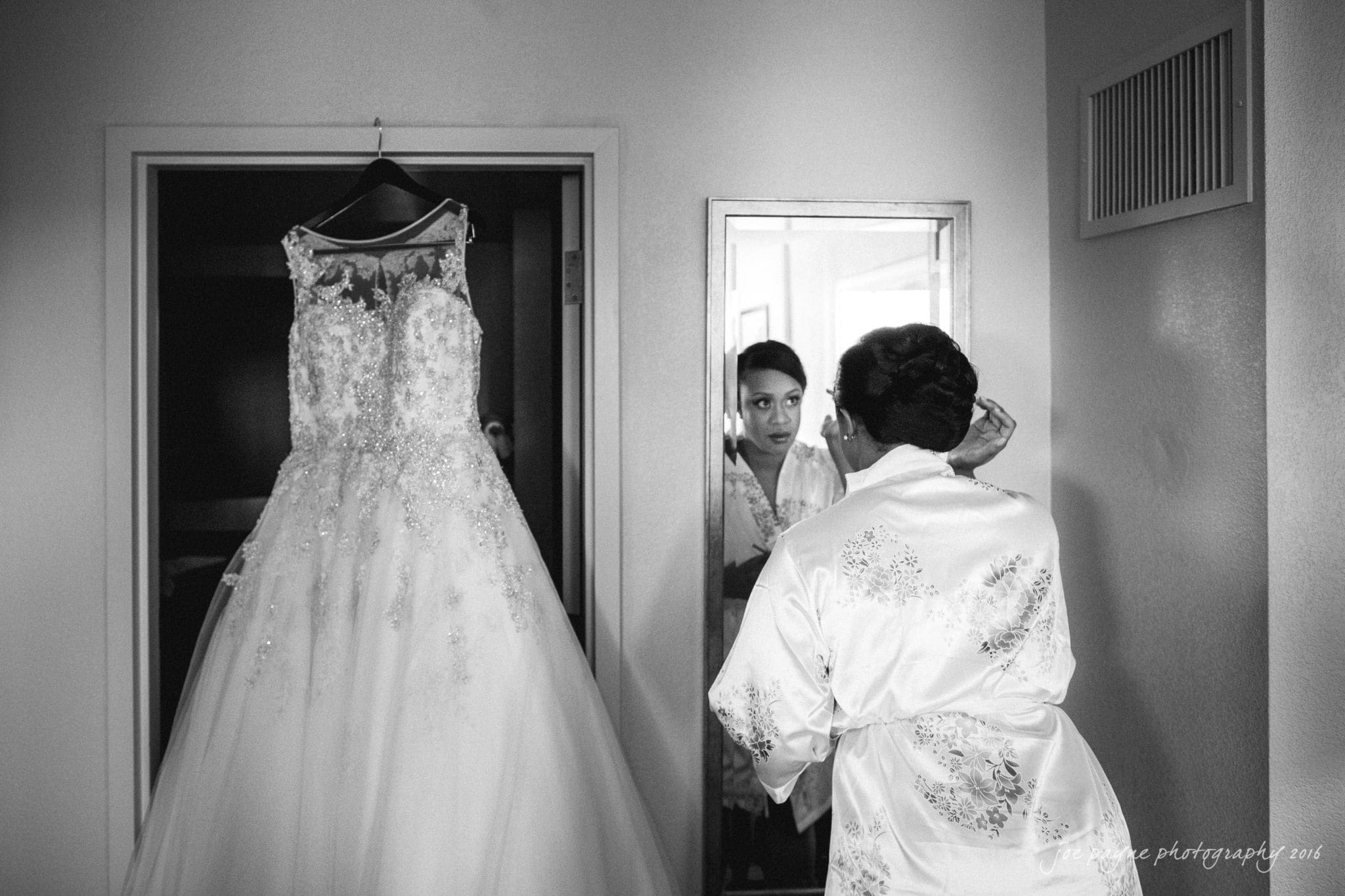 winston-salem nc wedding photography - toni & kevin A bride in a silk robe looks into a mirror, adjusting her hair. A lace wedding gown hangs nearby. The scene is set in a room with minimalist decor, capturing a serene moment as she prepares for her wedding day.