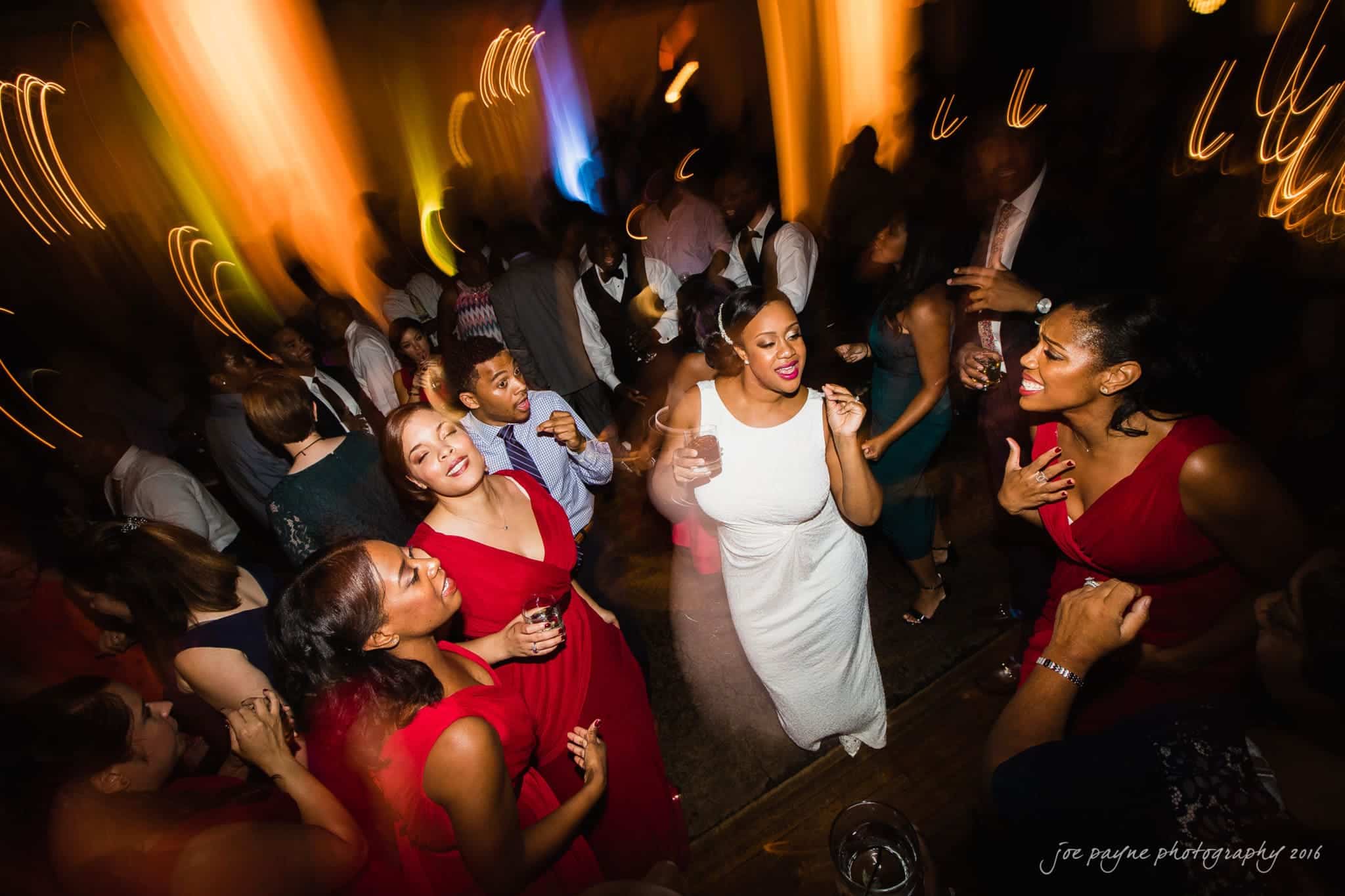 winston-salem nc wedding photography - toni & kevin A lively wedding reception with people dancing energetically. Guests are wearing formal attire; some women are in red dresses, and one is in a white dress. The room is warmly lit, creating a festive atmosphere with blurred movement effects.