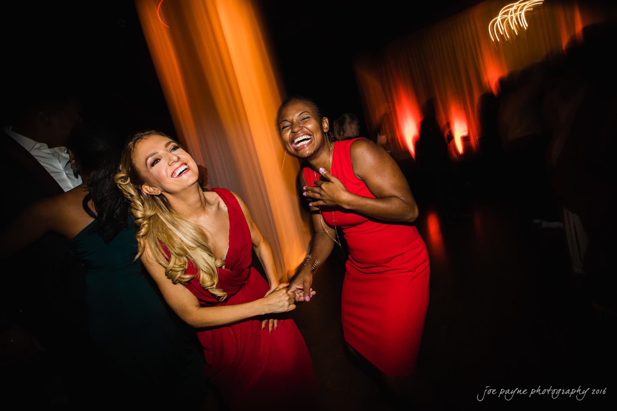 winston-salem nc wedding photography - toni & kevin Two women wearing red dresses are dancing and laughing on a dimly lit dance floor. Warm, blurred lights create a festive atmosphere in the background.