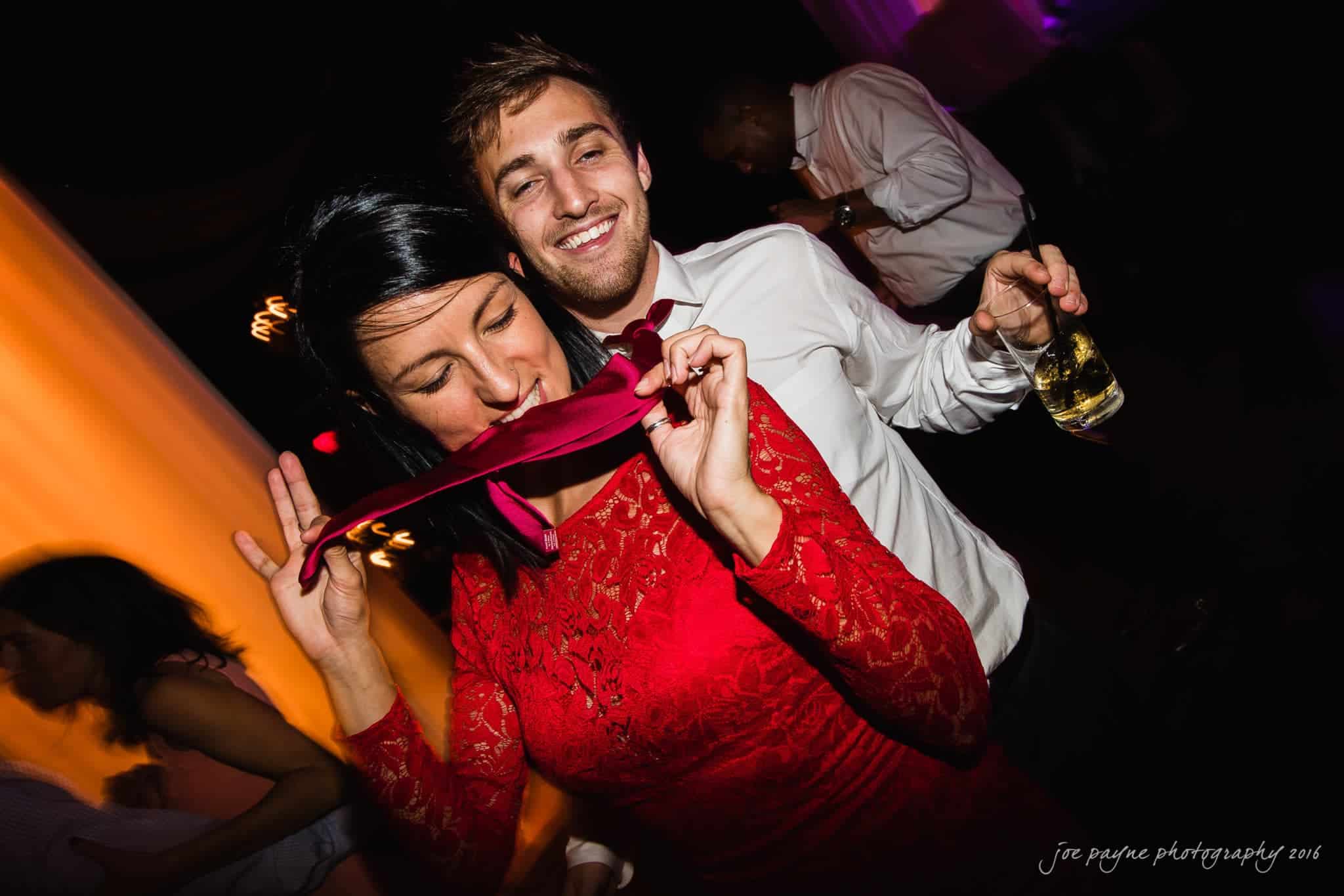 winston-salem nc wedding photography - toni & kevin A woman in a red dress playfully bites a necktie while a man in a white shirt smiles behind her. He holds a drink. They are at a lively party with people dancing in the background.