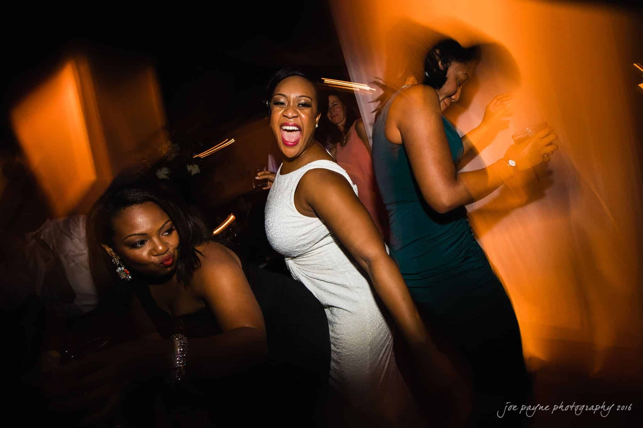 winston-salem nc wedding photography - toni & kevin A group of people dancing energetically in a dimly lit room. The woman in the center is wearing a white dress and smiling joyfully. Motion blur adds a sense of liveliness to the scene.