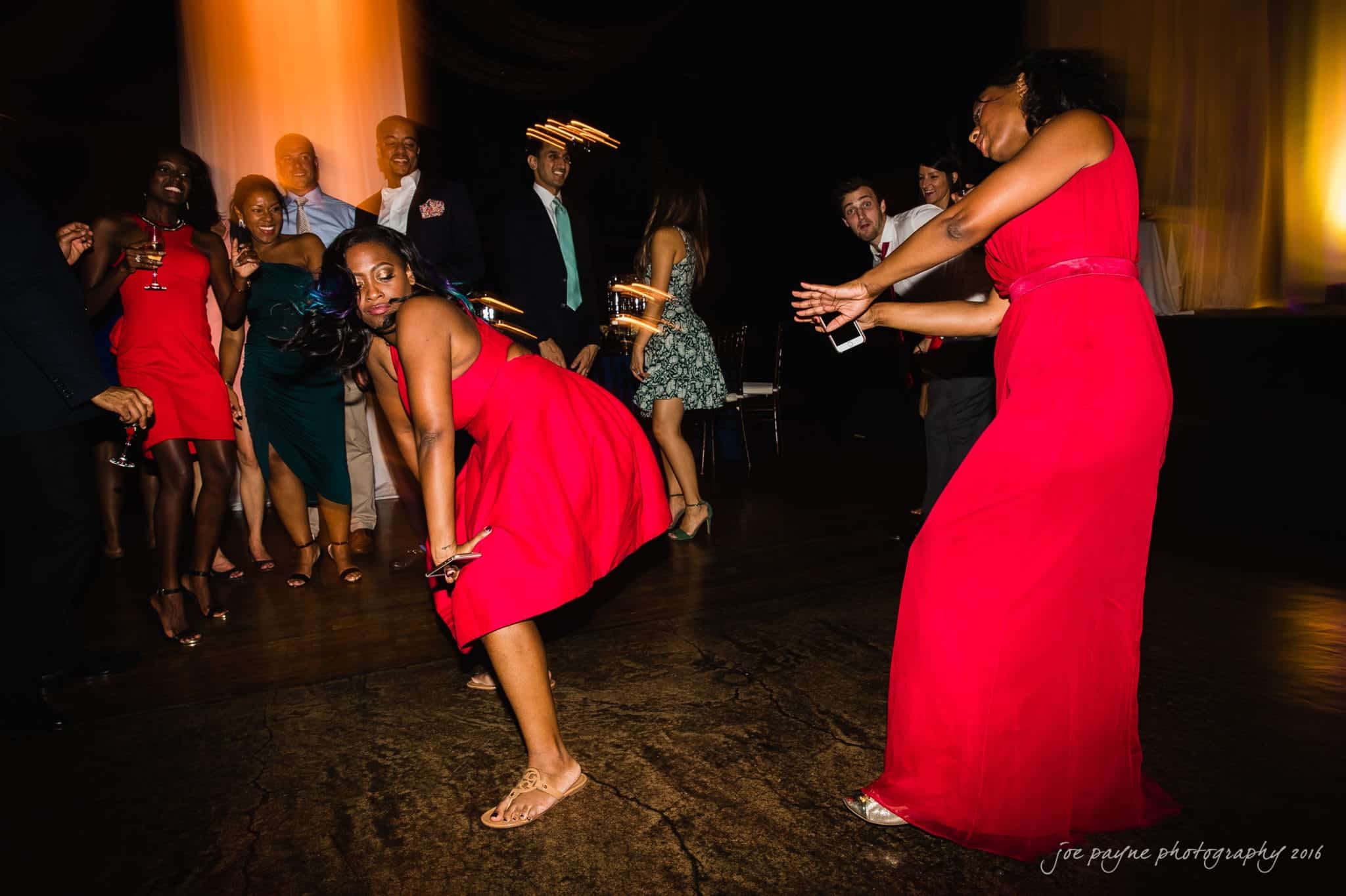 winston-salem nc wedding photography - toni & kevin A group of people in formal attire dance energetically at a party. Two women in red dresses are at the forefront, one crouched low and laughing. Others in the background are cheering or dancing. The atmosphere appears lively and joyful.
