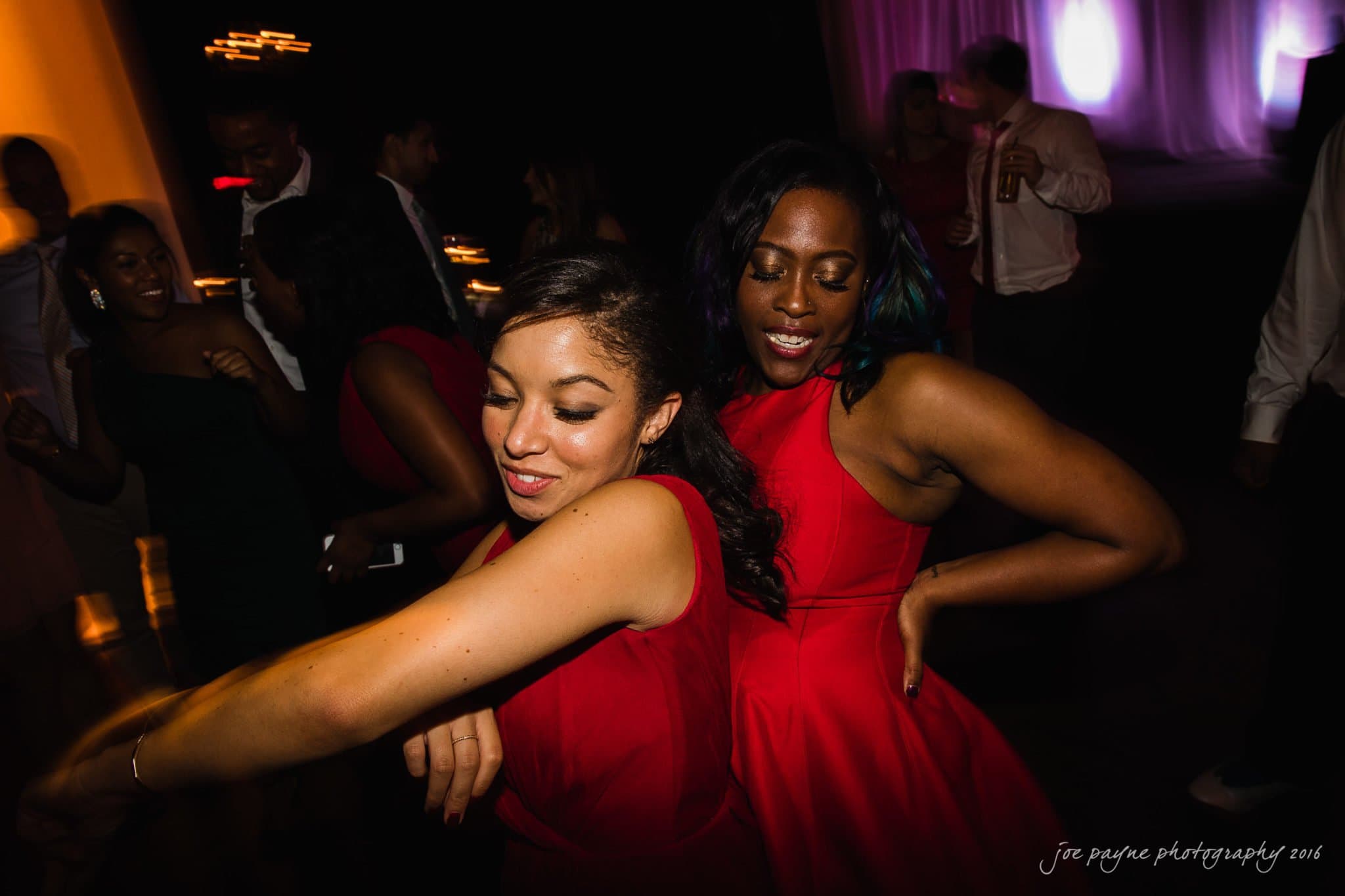 winston-salem nc wedding photography - toni & kevin Two women in red dresses are dancing together at a dimly lit event or party. One woman is smiling with her eyes closed, while the other has her hand on her hip. People in the background are chatting and holding drinks.