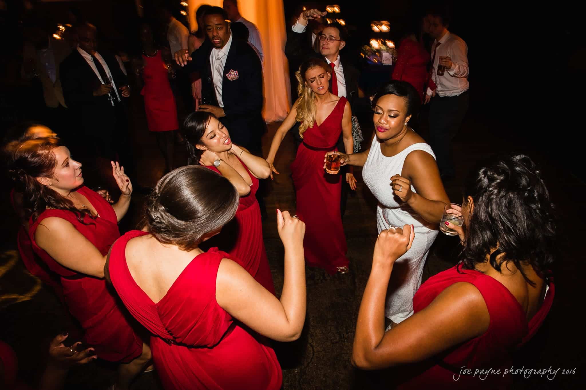 winston-salem nc wedding photography - toni & kevin A group of people in formal attire are dancing in a dimly lit setting. Several women in red dresses and a woman in a white dress are in the foreground, enjoying the music. Other attendees are visible in the background.