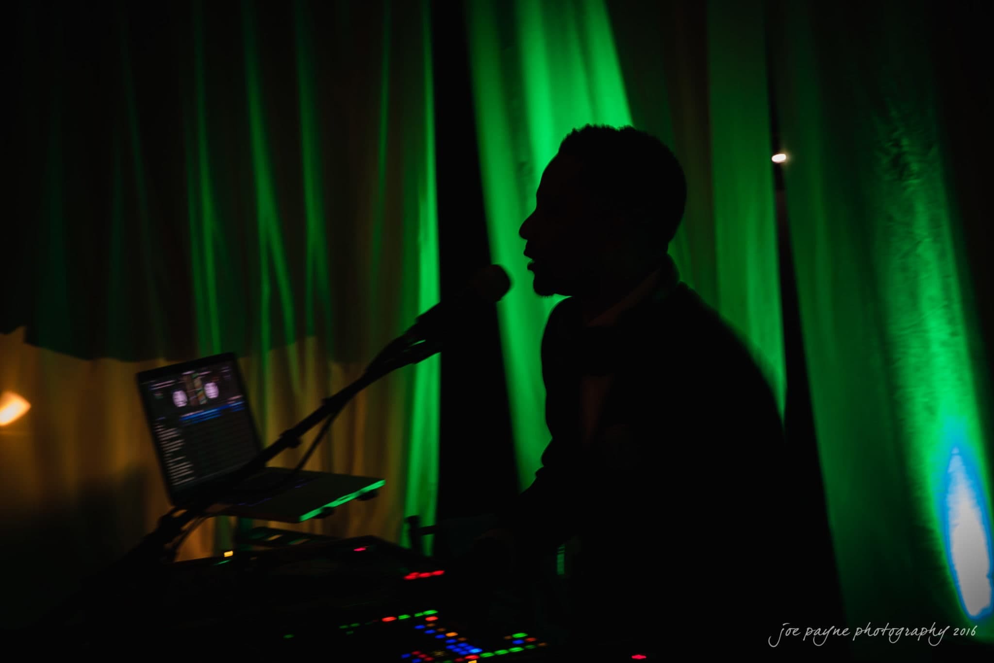 winston-salem nc wedding photography - toni & kevin Silhouette of a person singing into a microphone near a DJ setup with a laptop. The background is illuminated with vibrant green and yellow lights, creating a dramatic contrast.