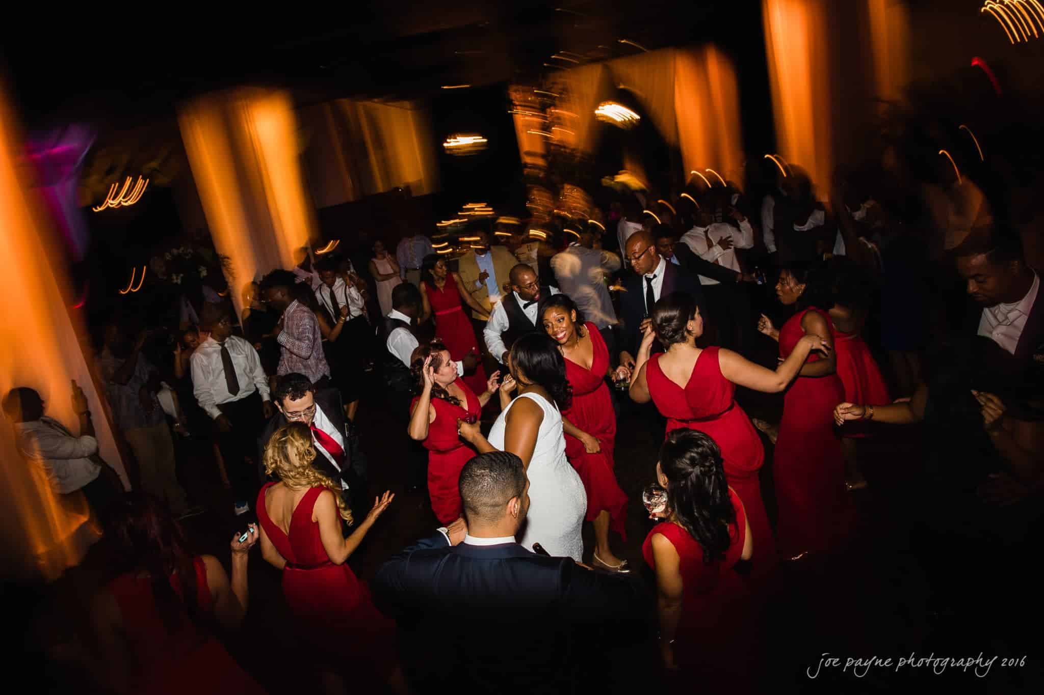 winston-salem nc wedding photography - toni & kevin A lively party scene with people dancing energetically in a dimly lit venue. Attendees are dressed in formal attire, with many wearing red dresses. The lighting creates dynamic, blurred motion effects.