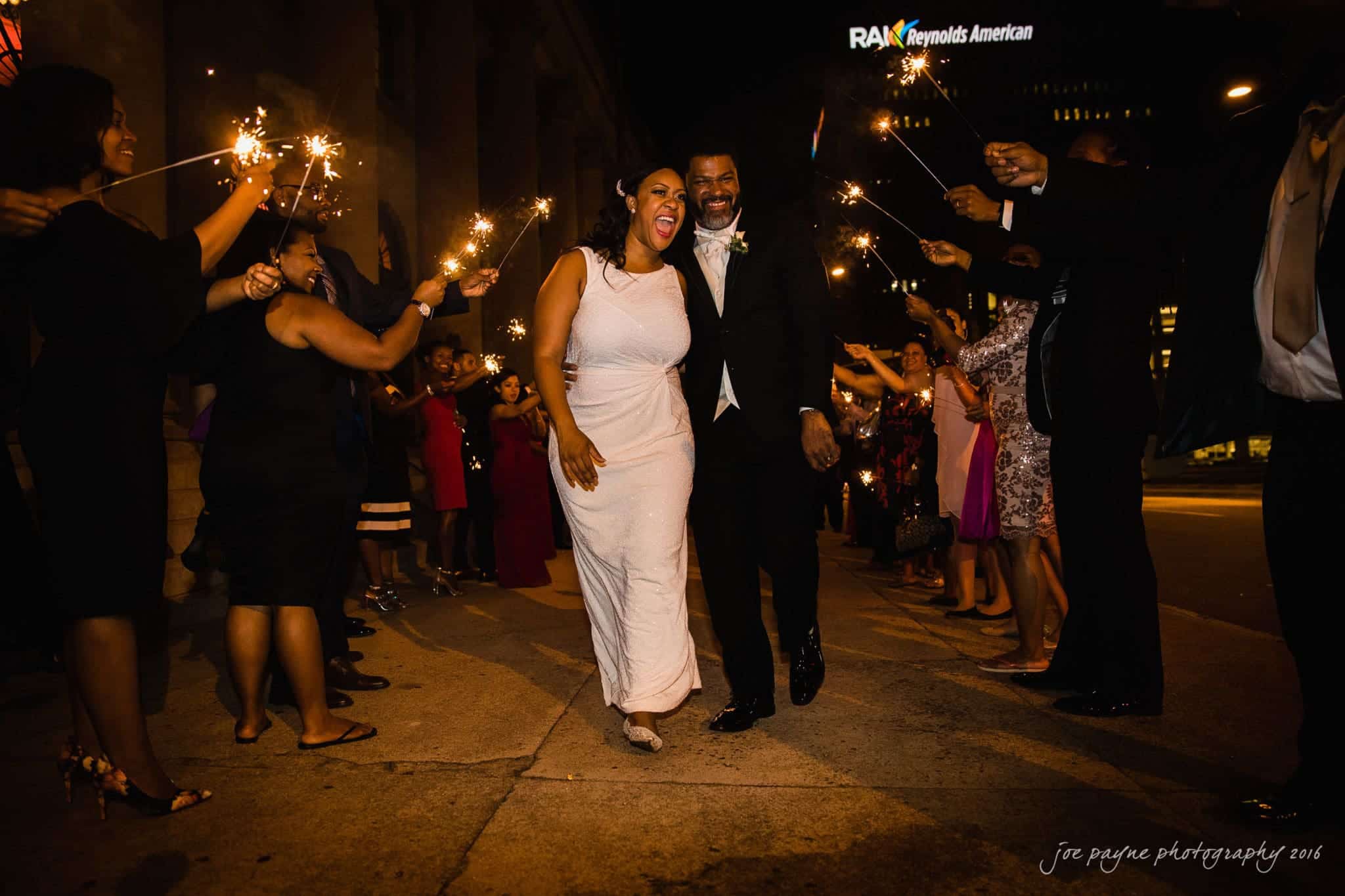 winston-salem nc wedding photography - toni & kevin A bride and groom in wedding attire walk joyfully through a crowd of people holding sparklers in a nighttime celebration. The crowd forms a tunnel with their sparklers, creating a festive and lively atmosphere.