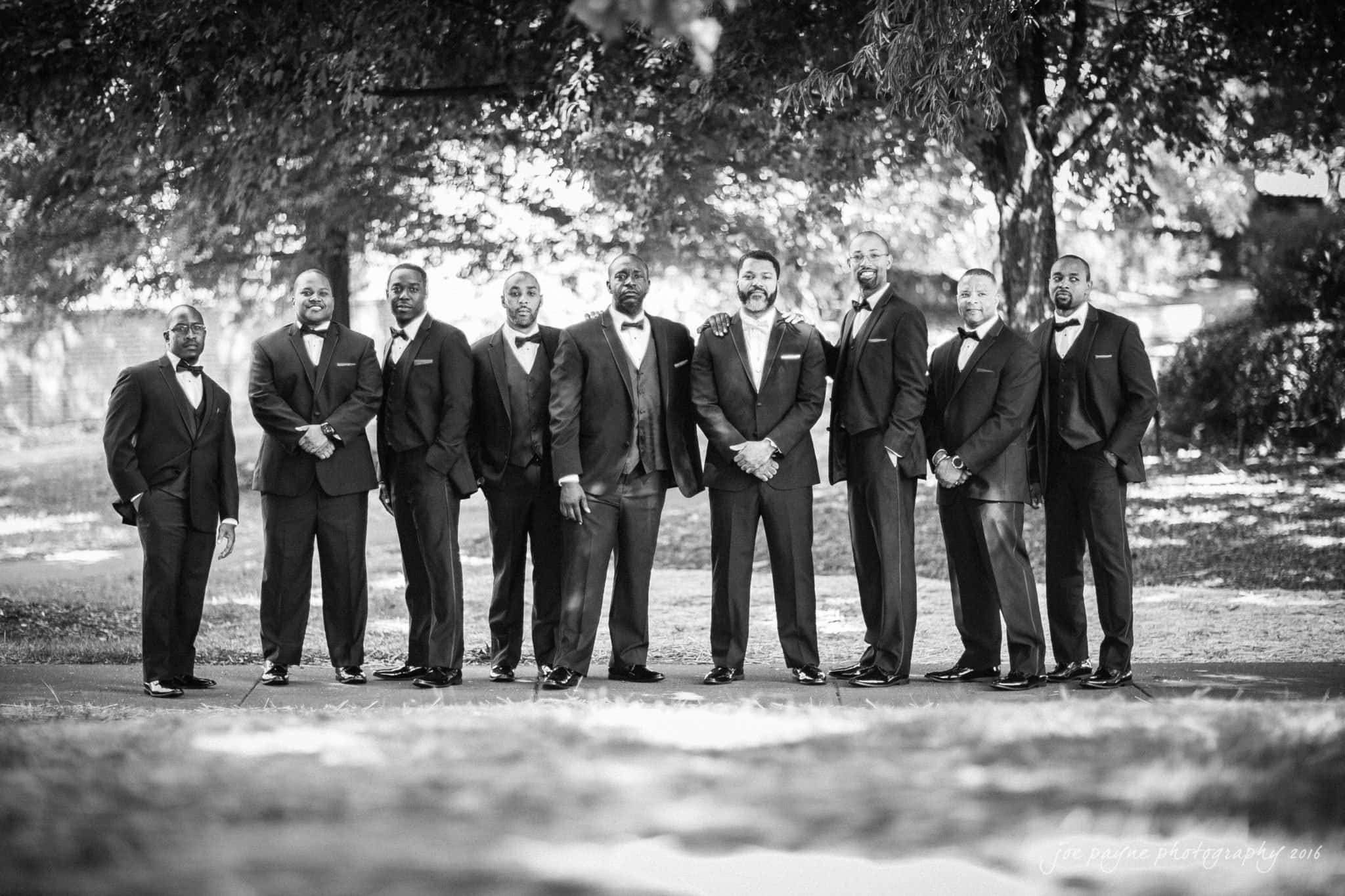 winston-salem nc wedding photography - toni & kevin A group of ten men in formal suits stand outdoors. They are arranged in a line, smiling at the camera, with trees and sunlight in the background. The black and white photo conveys a sense of elegance and camaraderie.