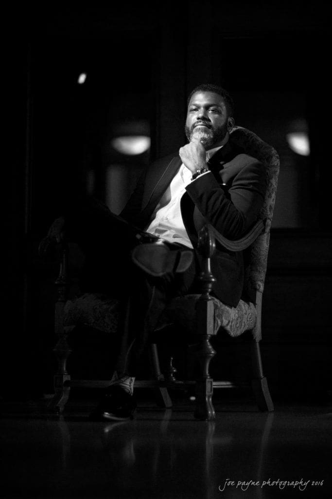 winston-salem nc wedding photography - toni & kevin A man in a suit sits confidently in an ornate chair, with one hand resting on his chin. The image is in black and white, creating a dramatic and contemplative atmosphere.