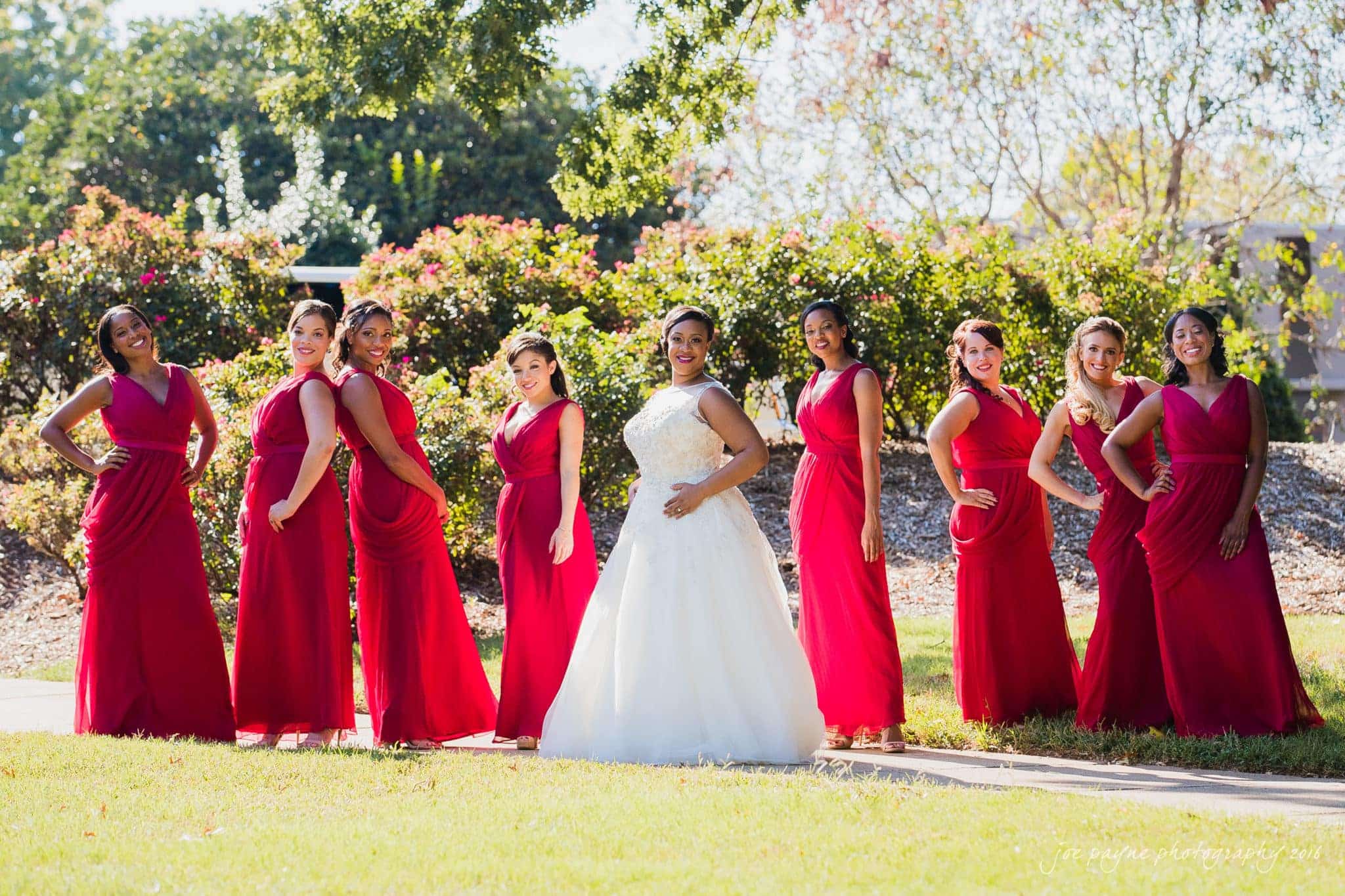 winston-salem nc wedding photography - toni & kevin A bride in a white dress poses outdoors with seven bridesmaids dressed in matching red gowns. They are all standing in a row, smiling, with greenery and trees in the background. The sun is shining brightly.
