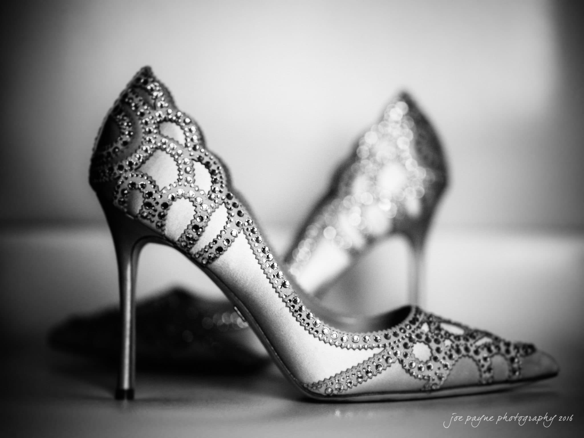 winston-salem nc wedding photography - toni & kevin A black and white image of elegant high-heeled shoes encrusted with intricate rhinestone patterns, positioned on a flat surface. The foreground shoe is sharply focused, while the background shoe is softly blurred, adding depth to the photo.