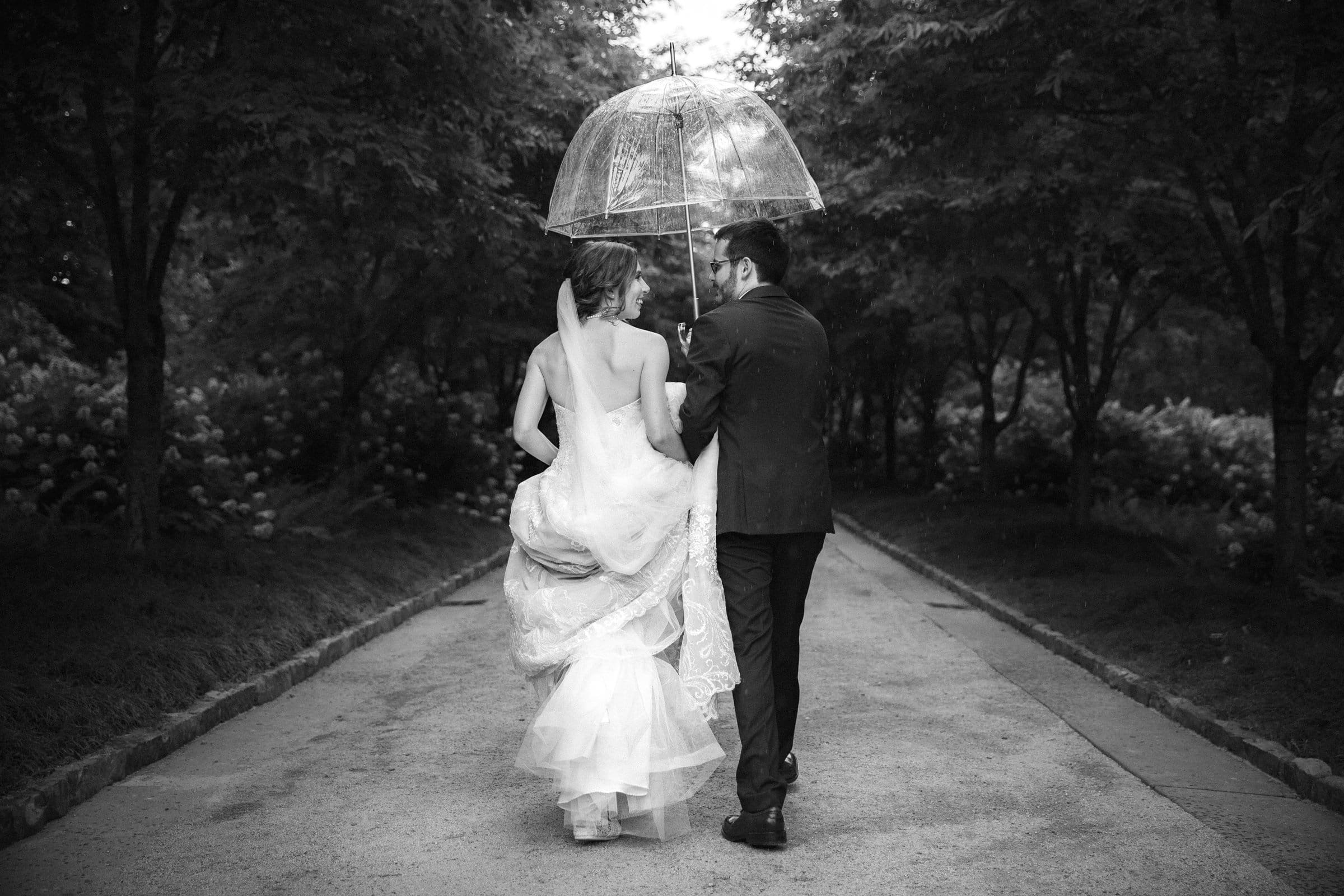 Duke Chapel Weddings A candid black and white wedding photograph of a bride and groom sharing an umbrella as they walk away down Cherry Allée at Sarah P. Duke Gardens, with the groom holding her dress tail as they talk.