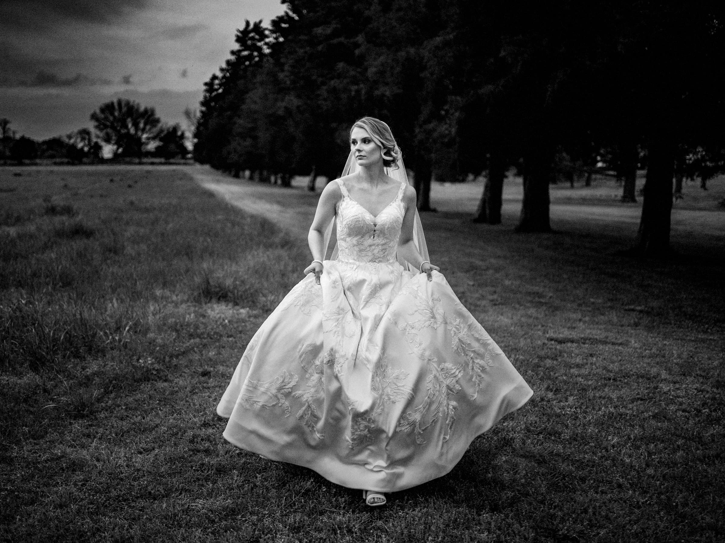 A black and white photo of a bride in an embellished wedding dress walking outdoors. She is holding the sides of her gown and gazing off to the side. The landscape features grass and a row of trees in the background.