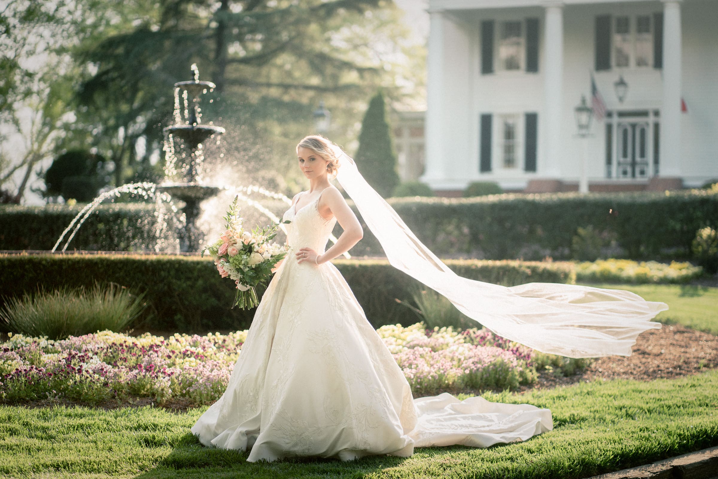 A bride in a flowing white gown stands gracefully in a garden, holding a bouquet of flowers. Her veil drapes behind her. A fountain and a large house with pillars are visible in the background. The scene is sunlit and serene.