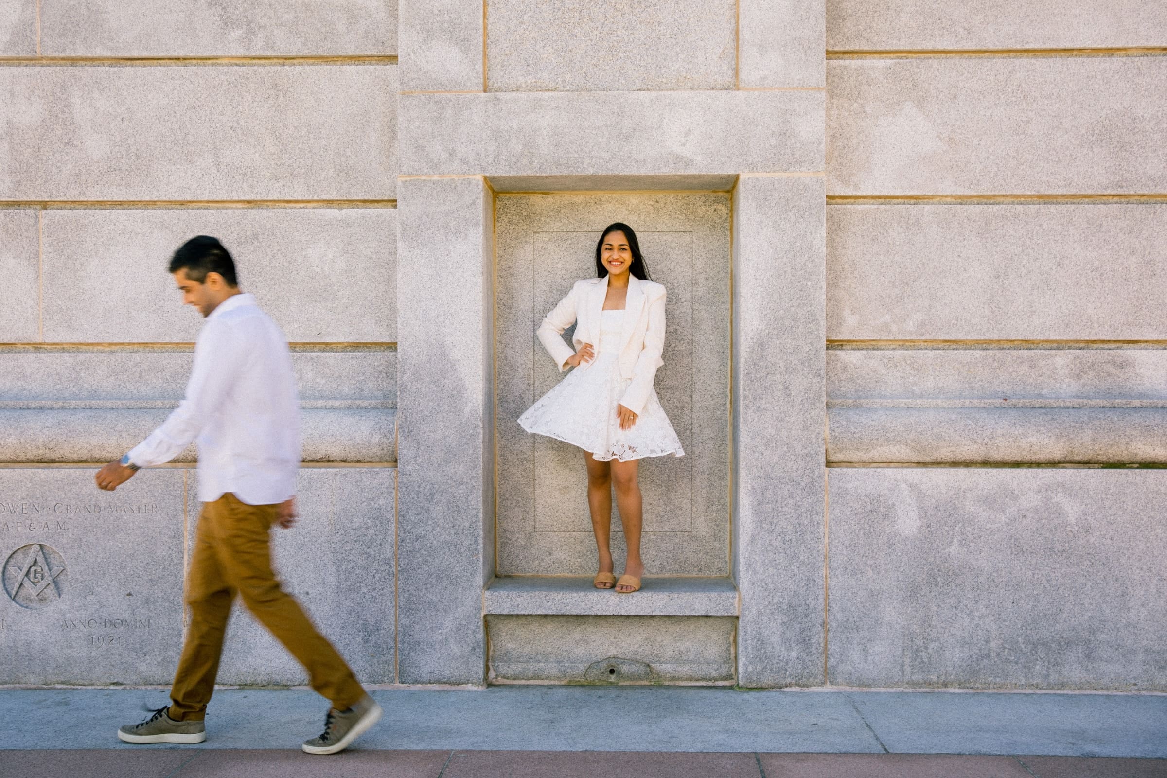 A woman in a white dress and blazer stands smiling in a small architectural alcove of a stone wall. A man in a white shirt and brown pants walks past in the foreground. The scene is outdoors on a sunny day.