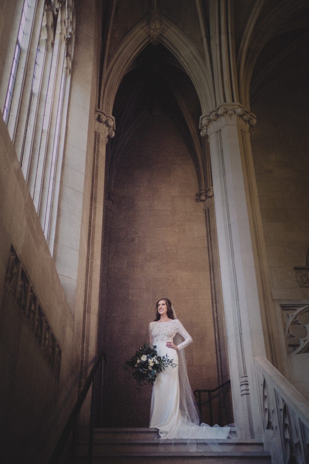 A striking bridal portrait inside Duke Chapel, featuring the bride standing beneath the towering gothic stone vaulted ceilings as she looks toward a massive, ornamental stained-glass window.