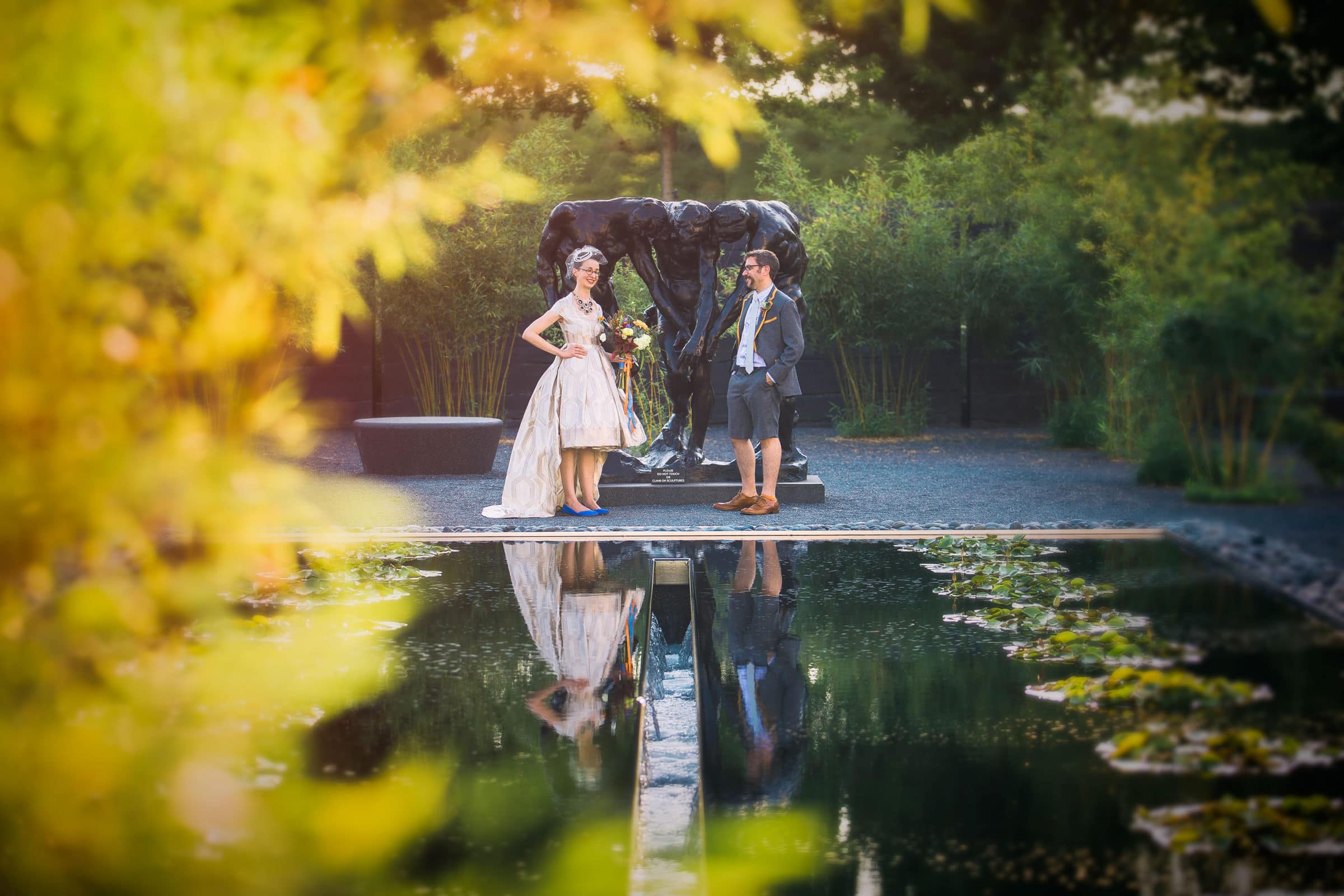A bride and groom stand facing each other by a reflective pond in a lush garden, their reflections mirrored in the water—a perfect moment beautifully captured by Raleigh NC wedding photographers.