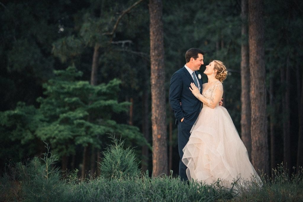 A bride and groom stand together in a forest, embracing and smiling at each other. The bride wears a flowing, light-colored gown, and the groom is in a dark suit. Raleigh NC wedding photographers capture this serene moment against the backdrop of tall trees.