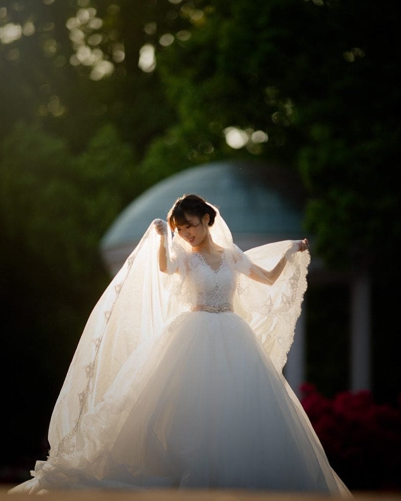 A bride in a white gown holds up her lace veil while standing outdoors. Sunlight filters through the trees, illuminating her dress. A gazebo is visible in the background amidst greenery.