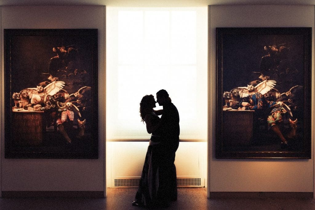 A silhouetted couple shares an intimate embrace—suggesting a moment of engagement—in front of a large bright window, framed by two dark, detailed paintings on either side, in a dimly lit gallery perfect for any romantic portfolio.