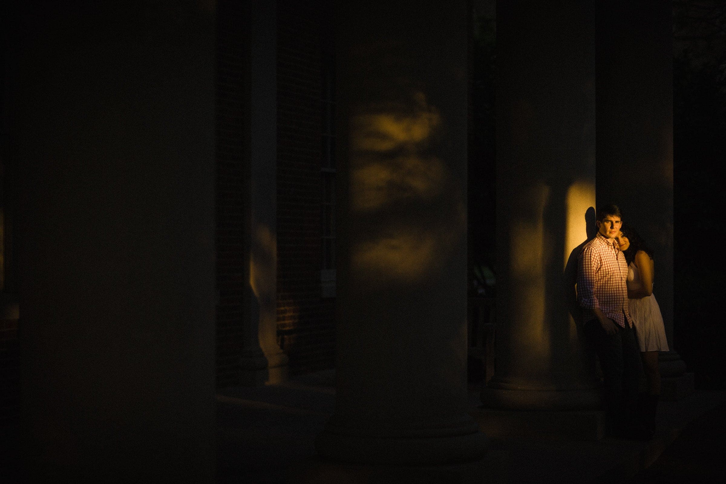 A man and woman stand close together between large columns, illuminated by a beam of warm light in an otherwise shadowy scene—an intimate moment captured perfectly for their Engagement Portfolio. The man faces forward as the woman gently leans on his shoulder.