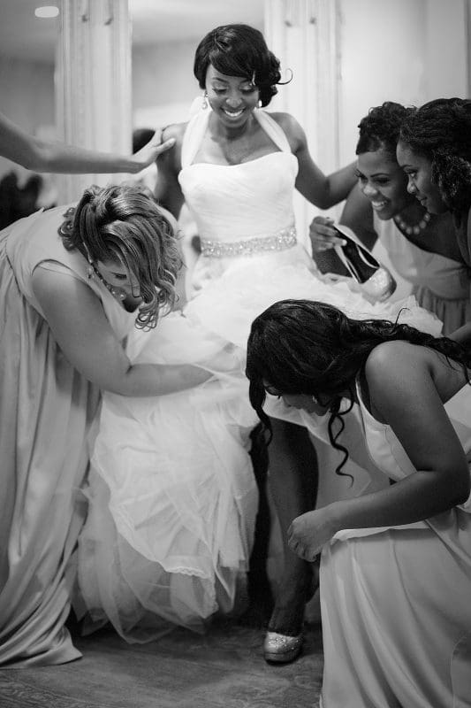 A bride in a white dress stands smiling as four bridesmaids adjust her gown. Captured by Raleigh NC wedding photographers, the scene is lively with bridesmaids focused on helping, creating a cheerful and supportive atmosphere. The image is beautifully captured in black and white.