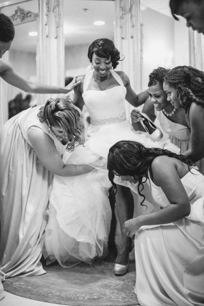 A bride in a white dress smiles as her bridesmaids, captured by Raleigh NC wedding photographers, help arrange her gown and shoes. They are gathered closely around her, laughing and adjusting the fabric in a joyful, supportive scene.