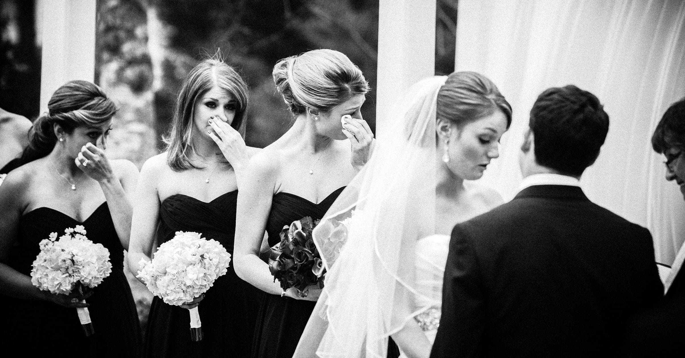 Bridesmaids in dark dresses wipe their tears while holding bouquets, standing behind a bride and groom during an emotional moment—perfectly captured by Raleigh NC wedding photographers.
