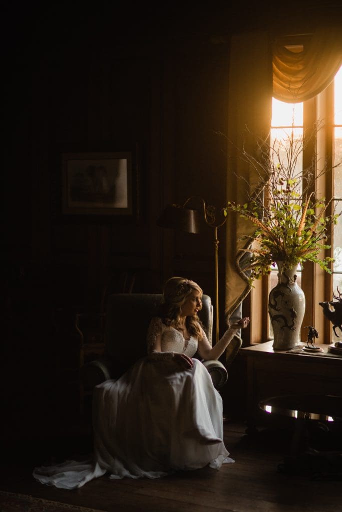 A woman in a white dress sits on a chair beside a window, bathed in warm light. She looks contemplatively at a large vase with flowers on a nearby table. The room is dimly lit, with dark wooden walls and curtains framing the window.