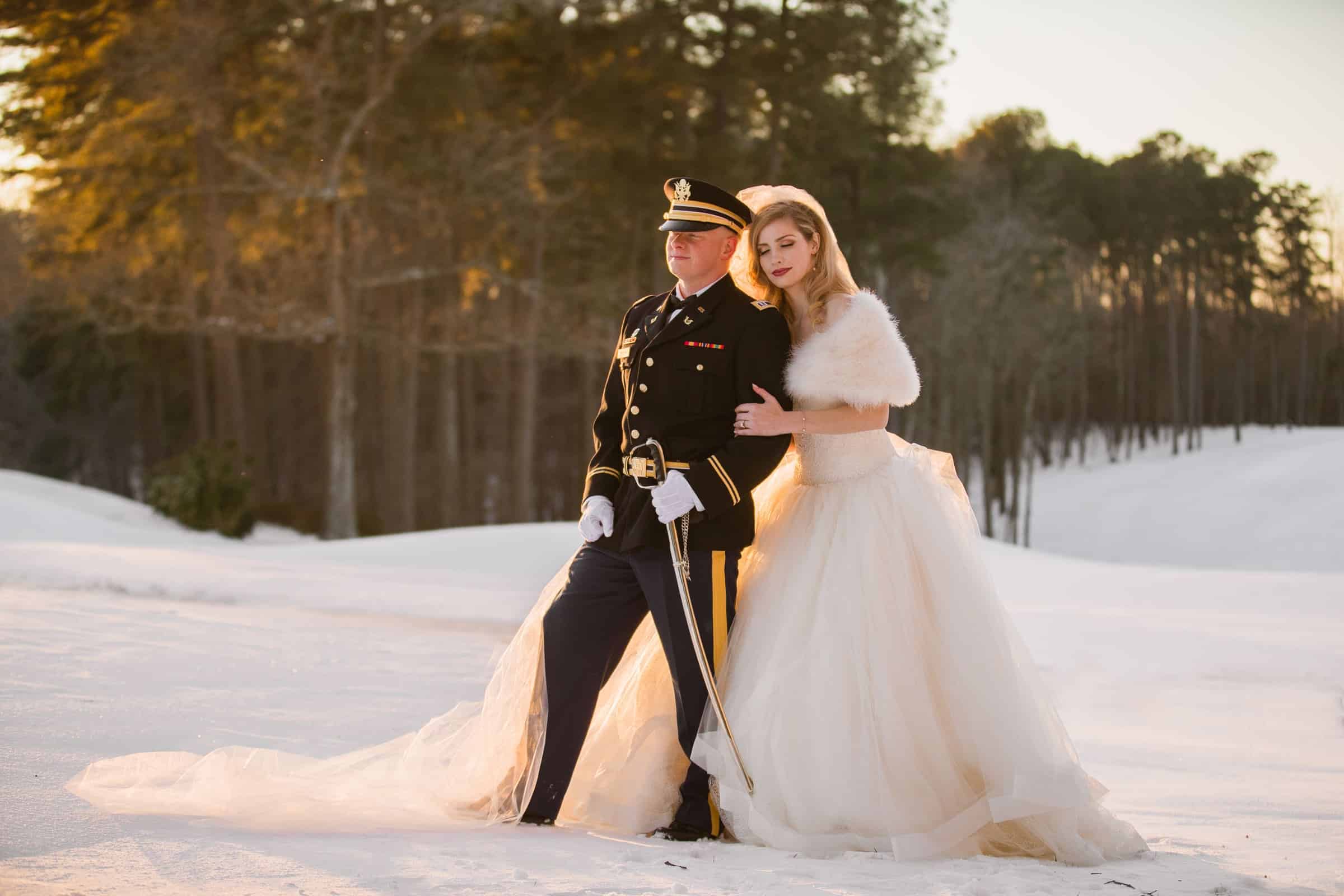 A bride in a white gown and fur stole stands arm-in-arm with a groom in a formal military uniform, posing together on snow-covered ground as Raleigh NC wedding photographers capture the moment among snowy trees.