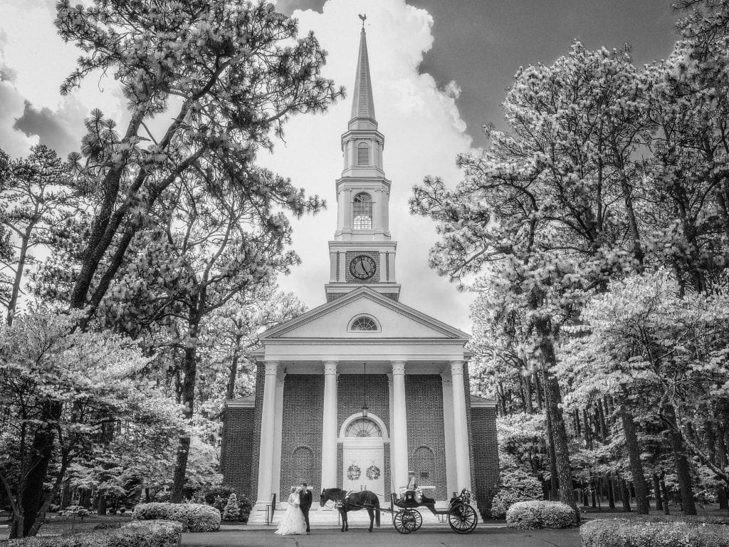 A black and white photo captures a timeless wedding scene in front of a church. A couple stands near a horse-drawn carriage, surrounded by trees, as dramatic clouds loom. With the touch of Raleigh NC wedding photographers, the image evokes elegance beneath the tall steeple and clock.