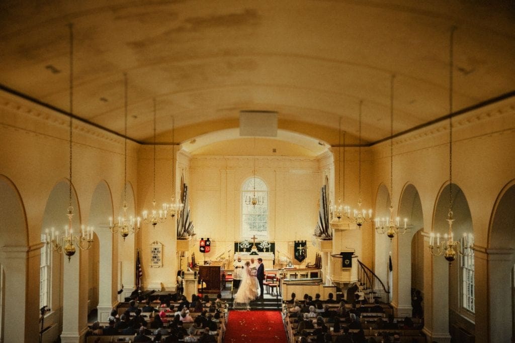 A panoramic view of a church interior during a wedding ceremony captured by Raleigh NC wedding photographers. The bride and groom stand at the altar, surrounded by guests seated in pews. The church is elegantly decorated with chandeliers and ornate arches.