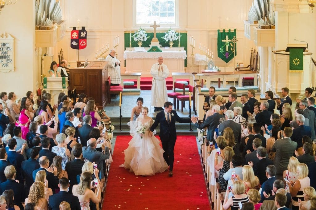 A newlywed couple walks down the aisle of the Village Chapel in Pinehurst, smiling and holding hands as Raleigh NC wedding photographers capture the moment. Guests stand and clap on either side, celebrating the occasion. The church is adorned with white flowers and green banners, while a priest stands at the altar in the background.