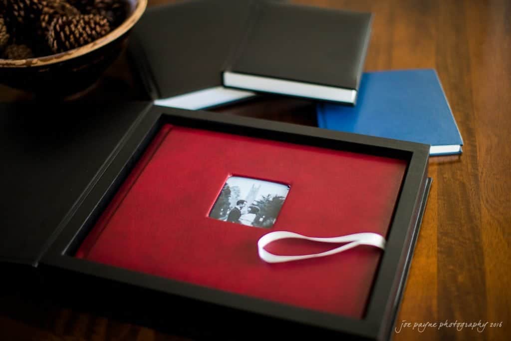 A red photo album with a small black-and-white photo on the cover sits open on a wooden table, showcasing Raleigh wedding memories. The album has a white ribbon bookmark. Nearby are closed black and blue albums, reminiscent of professional photographer collections, and a bowl of pine cones.