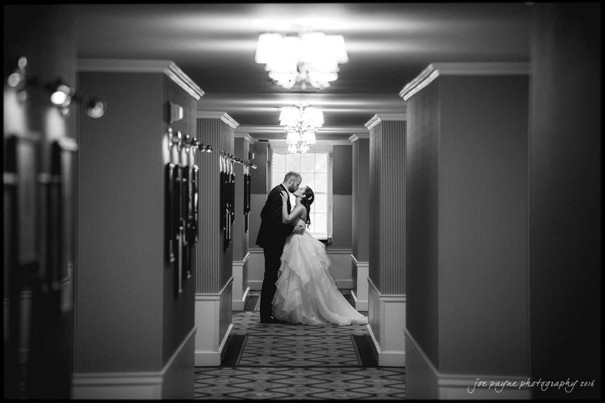 A bride and groom share a romantic moment in a dimly lit, elegant hotel hallway. The groom leans down to kiss the bride as she holds onto him, both in formal wedding attire. Photographs line the walls, and light fixtures hang from the ceiling.
