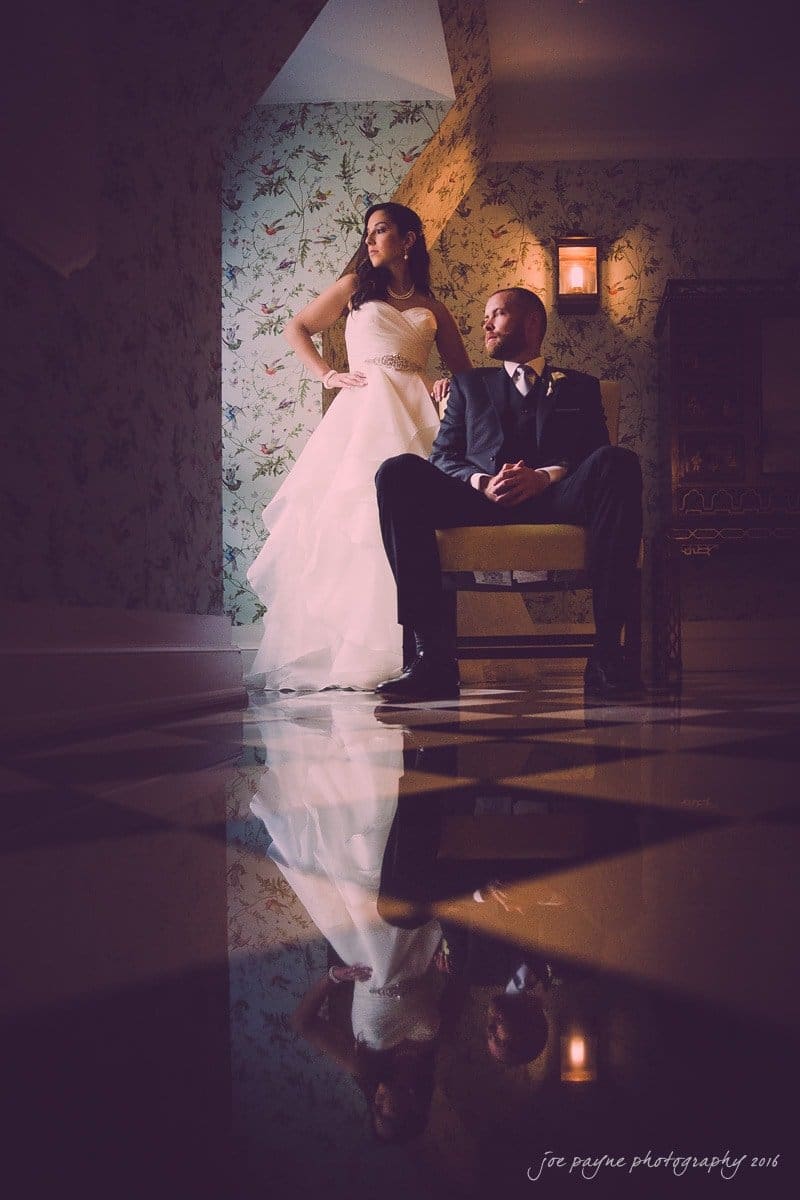 carolina inn wedding photography - kristen & matthew (x2) A bride in a white gown stands confidently next to a seated groom in a suit. They pose stylishly in a dimly lit room with floral wallpaper and a reflective floor, creating a dramatic atmosphere.