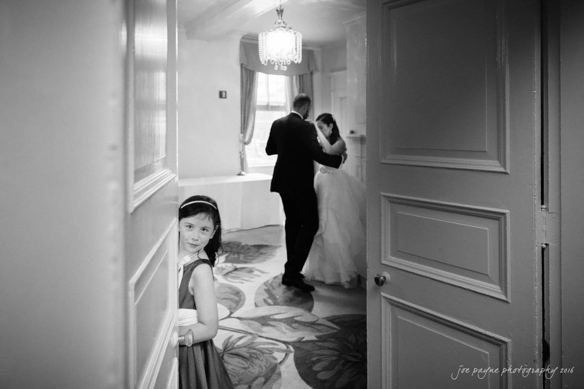 carolina inn wedding photography - kristen & matthew (x2) A young girl peeks around a door, watching a couple dance in a softly lit room. The scene has an intimate and joyful atmosphere, with a chandelier overhead and light streaming in through a window.