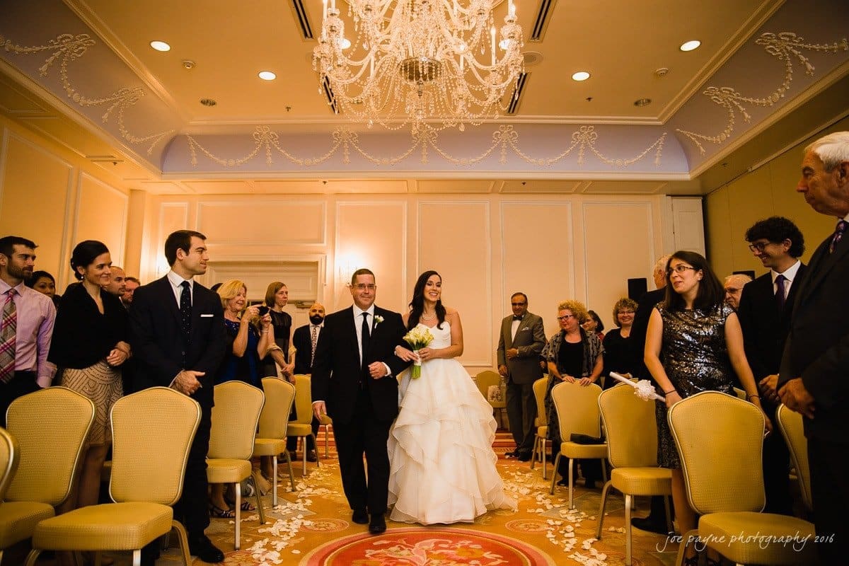 carolina inn wedding photography - kristen & matthew (x2) A bride in a white gown walks down the aisle with a man in a suit in a decorated indoor venue. Guests on both sides stand and smile, surrounded by elegant chandeliers and floral arrangements on the floor.